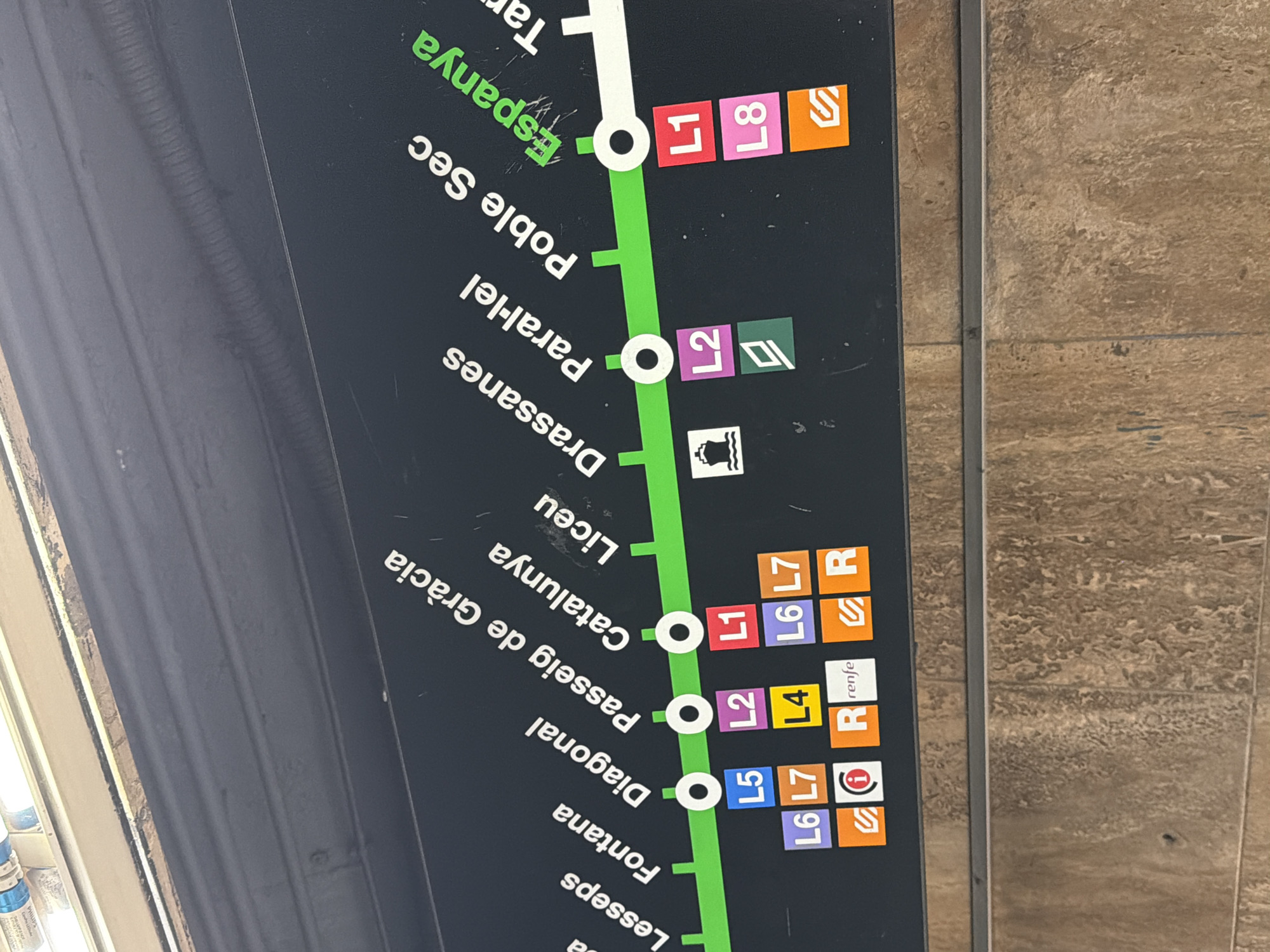 The image shows a section of a Barcelona metro map, indicating train lines and station names. The map features a dark background with green and white lines representing the metro routes. Station names such as "Lesseps", "Fontana", "Diagonal", "Passeig de Gràcia", "Catalunya", "Liceu", "Drassanes", "Paral·lel", "Poble Sec", and "Espanya" are displayed in white text, angled upwards to the left. Below some of these station names are colored squares with numbers and symbols, indicating different metro lines (L1, L2, L4, L5, L6, L7, L8) and other transport services, such as the Renfe train network (indicated by "R renfe"). The map appears to be a public display, likely located within a metro station.
While the map itself is for the Barcelona metro system, the prompt asks to include the location in the city of l'Hospitalet de Llobregat, Spain. L'Hospitalet de Llobregat is a municipality bordering Barcelona and is served by several lines of the Barcelona Metro, including some that are visible on this map (e.g., L1 and L5 pass through stations in L'Hospitalet). Therefore, this map section could be found in a metro station within l'Hospitalet de Llobregat, or a station in Barcelona that serves as an interchange for lines extending into l'Hospitalet.
The image is a close-up, slightly angled view of the map. The environment appears to be indoors, with artificial lighting. There are no people or specific actions visible in the frame, only the static display of the map. The lower part of the image shows a textured surface, possibly a wall or floor, in earthy tones. The overall condition of the map display suggests it is in use, with some minor scuff marks visible on the dark background.