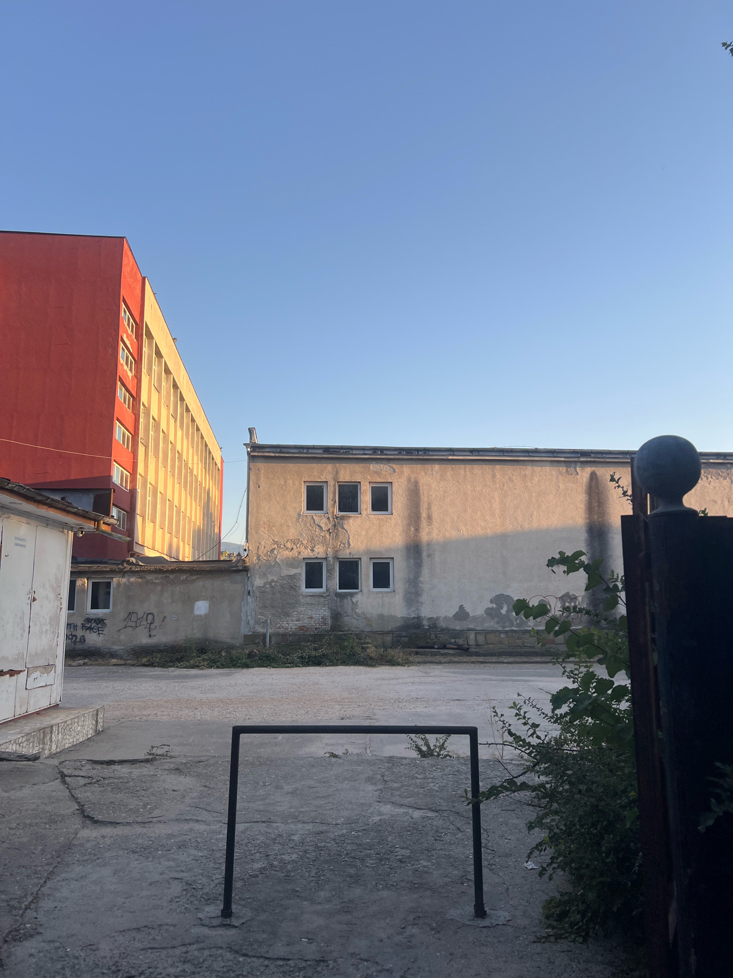 The image depicts an urban scene with two buildings and an empty outdoor space. On the left, a tall, multi-story building with a red facade is partially visible, with its yellow-toned upper floors featuring rows of windows. Adjacent to it, to the right, is a lower, weathered concrete building with several windows. Graffiti is present on the walls of the structures and the ground. The foreground shows a cracked concrete surface with a simple black metal frame structure standing in the center. To the far right, a dark metal fence post topped with a spherical ornament is visible, along with some greenery. The sky is clear and blue, suggesting daytime.
This image was taken in Plovdiv, Bulgaria, specifically in the area of the former "Balkan" factory, now known as the "Kapana Creative District" or "Tsar Simeon's Garden" area, which has seen redevelopment. The architecture and state of the buildings, including the graffiti, are characteristic of post-industrial or transitional urban environments in cities like Plovdiv.