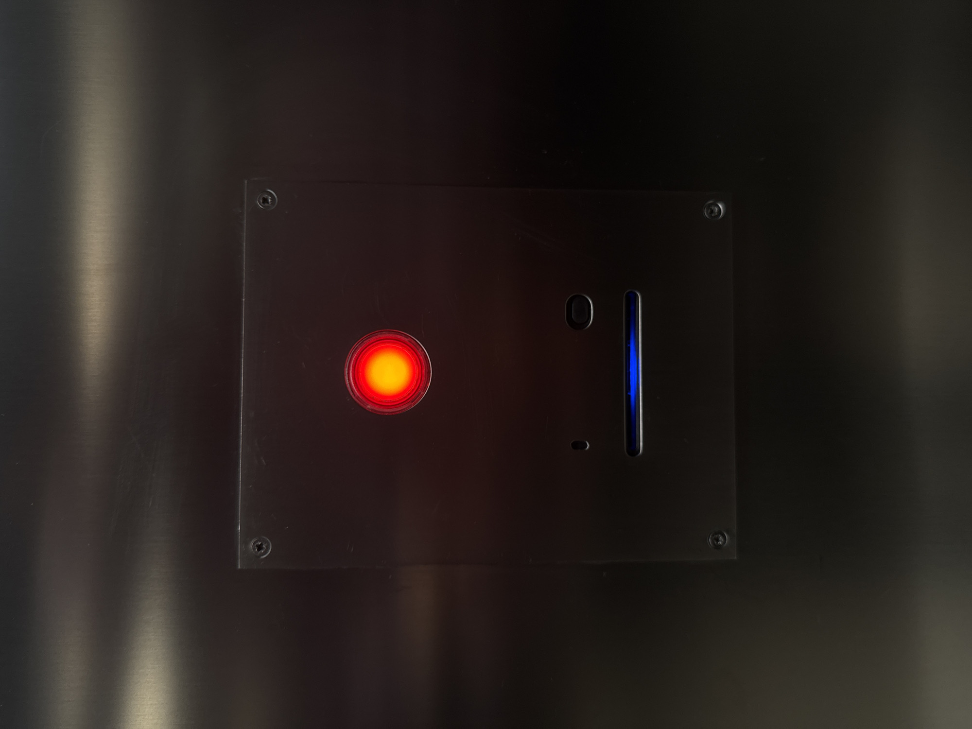 The image depicts a close-up of a metal panel with various illuminated features. On the left, there is a prominent red circular light. To the right, there is a slot-like aperture emitting blue light, with a small circular indent near it. The panel is secured by screws in each of its four corners, embedding it into a darker metal wall. There is no indication that this image is taken in Rome, Italy.