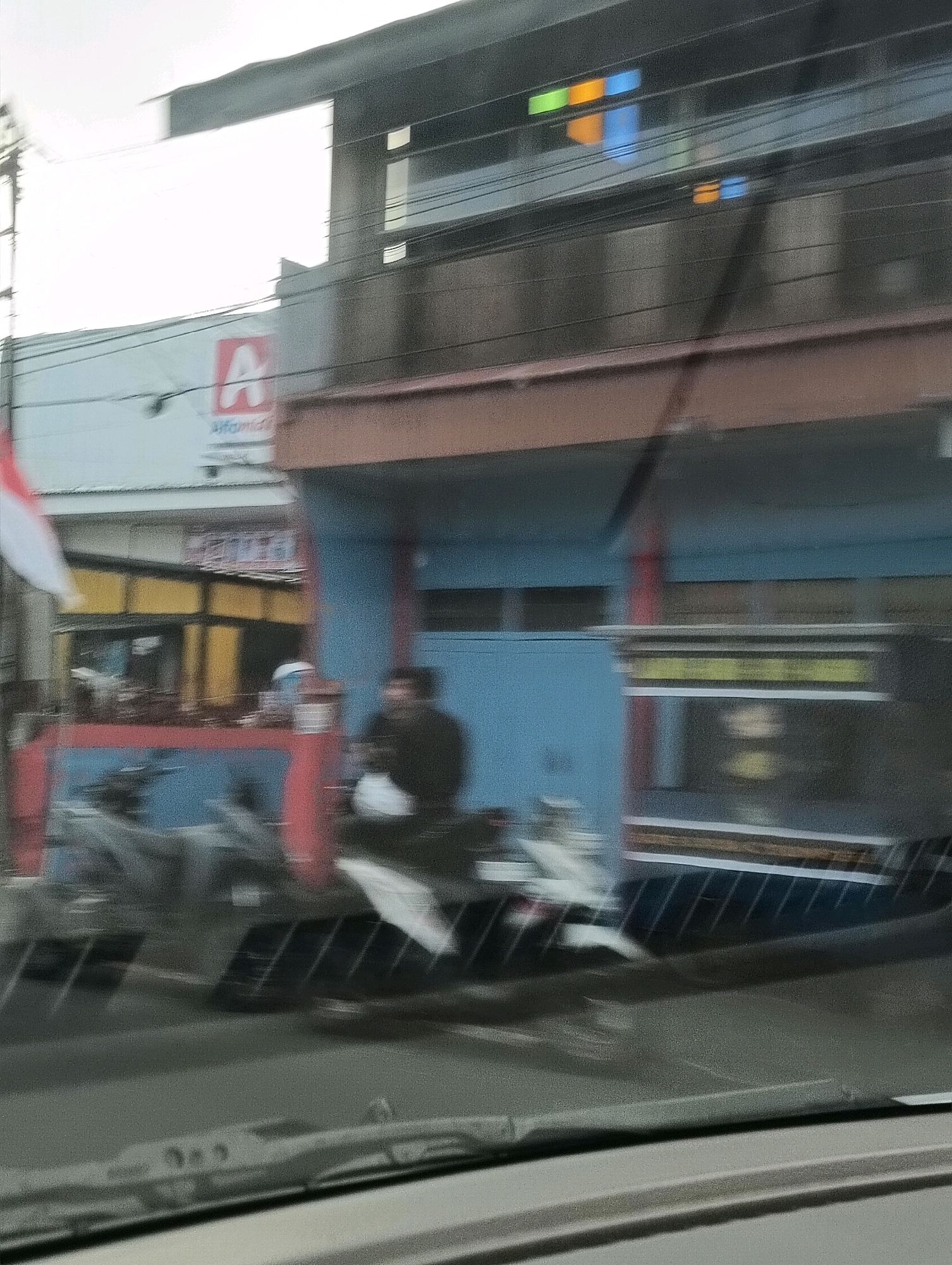 The image depicts a street scene in Tomohon, Indonesia, captured from inside a vehicle, as evidenced by the windshield wipers in the foreground and the motion blur. The focal point is a building with a blue facade and a brown overhang. To the left of the building, a red and white Indonesian flag is visible. A sign with a red 'A' logo and text that appears to be "Alfamart" is mounted on the building. Below the sign, a yellow storefront with dark framing is visible. In front of the building, two people on motorcycles are present. One person is wearing dark clothing and appears to be holding a white bag. The other motorcycle is mostly obscured. The street is paved and there are power lines overhead. The overall lighting suggests it is daytime, possibly overcast.
