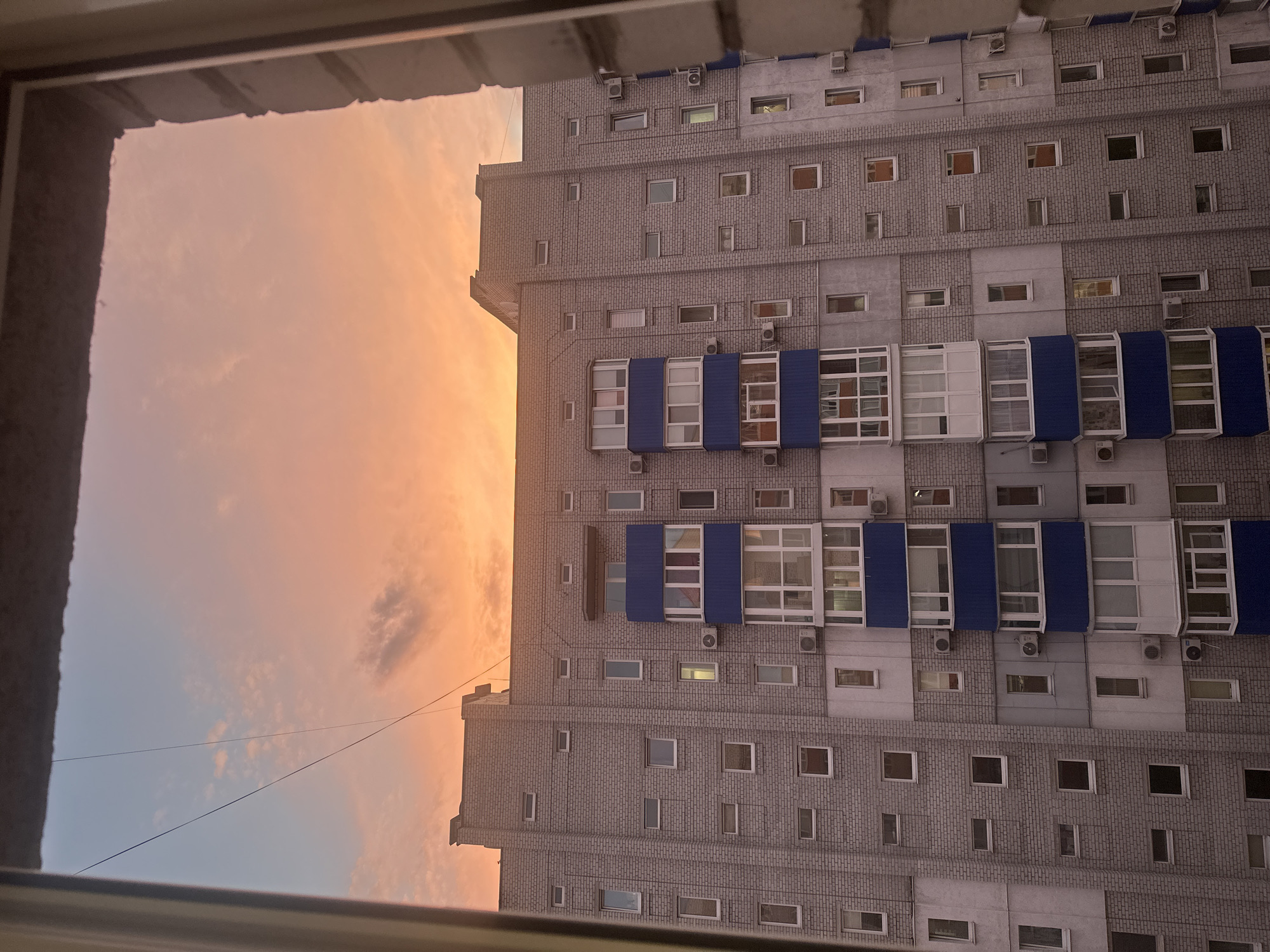 This is a vertical, eye-level, wide shot of a modern apartment building at sunset. The building dominates the right two-thirds of the frame, with a dramatic sunset sky filling the left third. The sky is a gradient of soft oranges, pinks, and blues, with wispy clouds. A thin wire stretches diagonally across the sky.
The apartment building appears to be constructed from grey brick with some concrete panels. It has a repetitive facade with numerous windows of varying sizes, many of which are dark, suggesting the apartments are not all lit. Some windows have air conditioning units mounted beneath them. A distinctive feature is a section of balconies or enclosed sunrooms with blue panels and large glass windows, located roughly in the middle of the building's visible height. The building is tall, with many stories, and its scale is emphasized by the perspective from below.
The time of day is clearly sunset, indicated by the warm colors of the sky. The weather appears clear, with only scattered clouds. The location is suggested to be Horishni Plavni, Ukraine. The overall mood is calm and perhaps a little melancholic, with the setting sun casting a serene glow over the urban landscape. There is no visible text in the image.