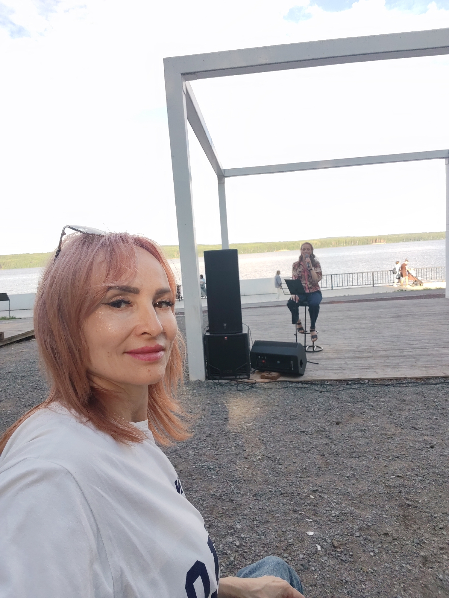 The media file is a selfie taken outdoors, with the focus on a woman in the foreground. She has reddish-pink hair, is wearing a white t-shirt with dark blue lettering, and has sunglasses on top of her head. She is smiling and looking towards the camera.
In the background, a stage is set up with wooden flooring. On the stage, a woman is seated on a stool, holding a microphone and appearing to be singing or speaking. There is a laptop in front of her and a set of speakers to her left. Further in the background, a body of water is visible, bordered by trees on the far side, suggesting a lakeside or riverside setting. The sky is overcast with patches of blue. The overall scene appears to be a casual outdoor performance or gathering. The time of day seems to be daytime, with soft lighting. There is no visible text that can be clearly read. The location is identified as Chaikovsky, Russia.