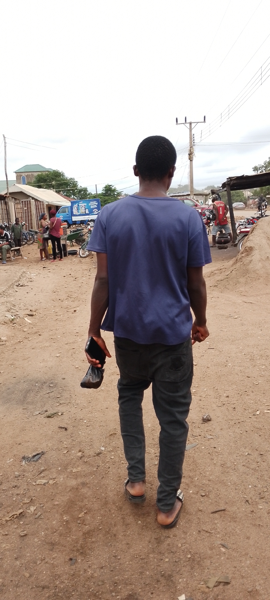 A full-shot, ground-level, eye-level, back-view, medium shot of a dark-skinned young male walking down a dusty, dirt road in Gboko, Nigeria. The young male is wearing a navy blue t-shirt, dark grey slim-fit pants, and black sandals with silver embellishments on the right sandal. He is holding a black cellphone in his right hand and a black plastic bag in his left hand. The road is unpaved, with loose dirt and scattered debris. In the background, there are several market stalls, motorcycles, a blue truck with "So Good Creamy" written on its side, a red car, and utility poles with power lines. The sky is overcast.