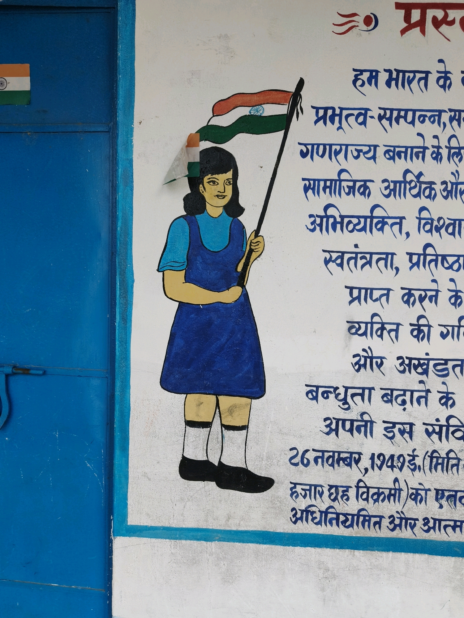 This is an image of a mural painted on a white wall, likely located in Katihar, India. The mural depicts a young girl holding a flag of India, with the Indian flag's colors of saffron, white, and green visible. The girl is wearing a blue dress and white socks with black shoes. To the left of the mural is a blue metal door with a visible latch. The wall itself appears to be textured and slightly aged. In the upper right corner of the mural, some text in Hindi is visible, which appears to be part of a preamble or pledge, possibly related to the Indian Constitution. The overall scene suggests a public space, perhaps a school or community building, decorated with patriotic imagery.