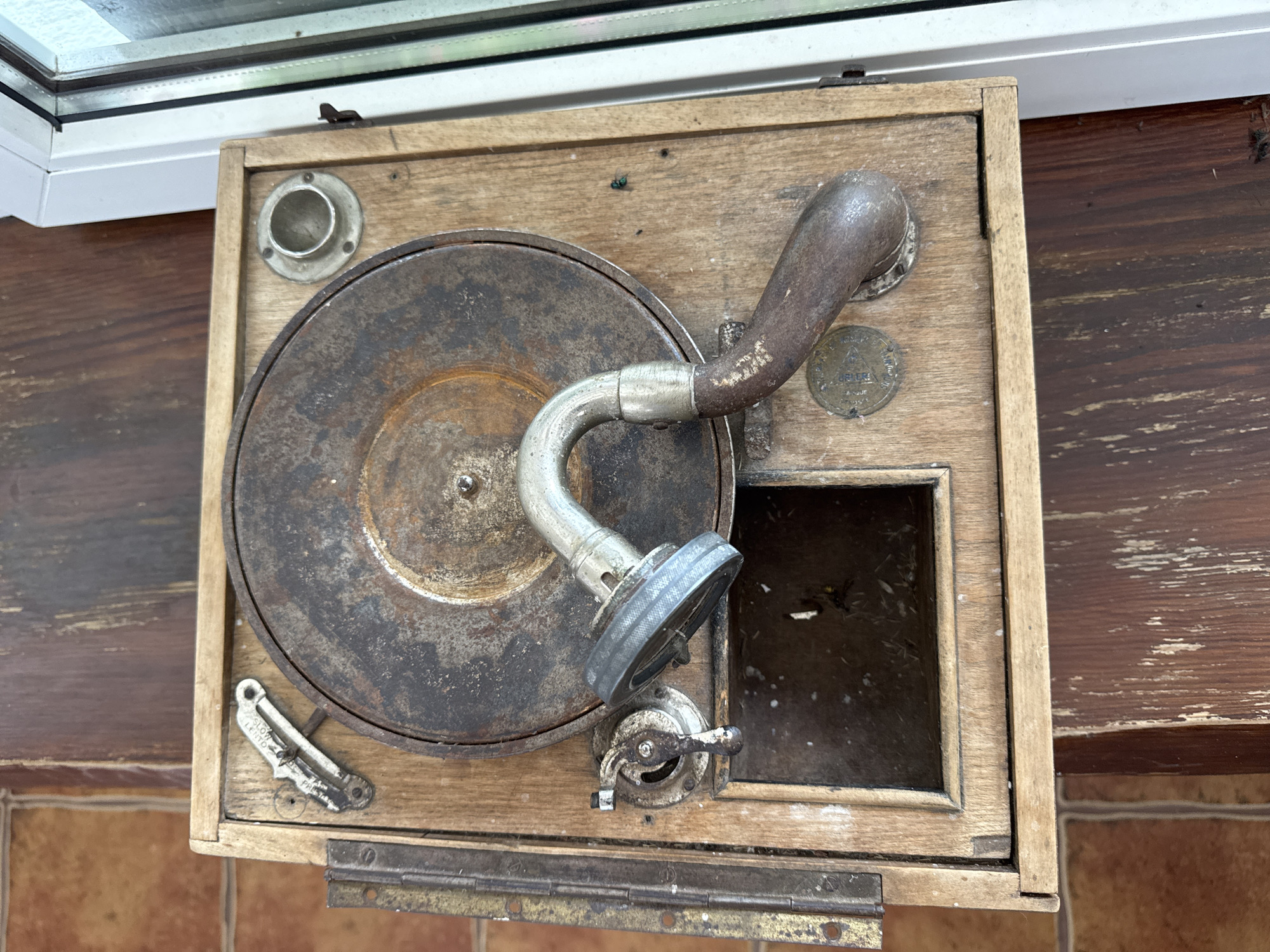 An antique gramophone sits on a wooden surface. The gramophone is made of wood and has a rusted metal turntable. A tone arm with a needle is positioned over the turntable. There is a small, open compartment on the gramophone's base. The gramophone is located on what appears to be a windowsill, with a view of greenery outside the window. The area is in Мірскі сельскі Савет, Belarus.