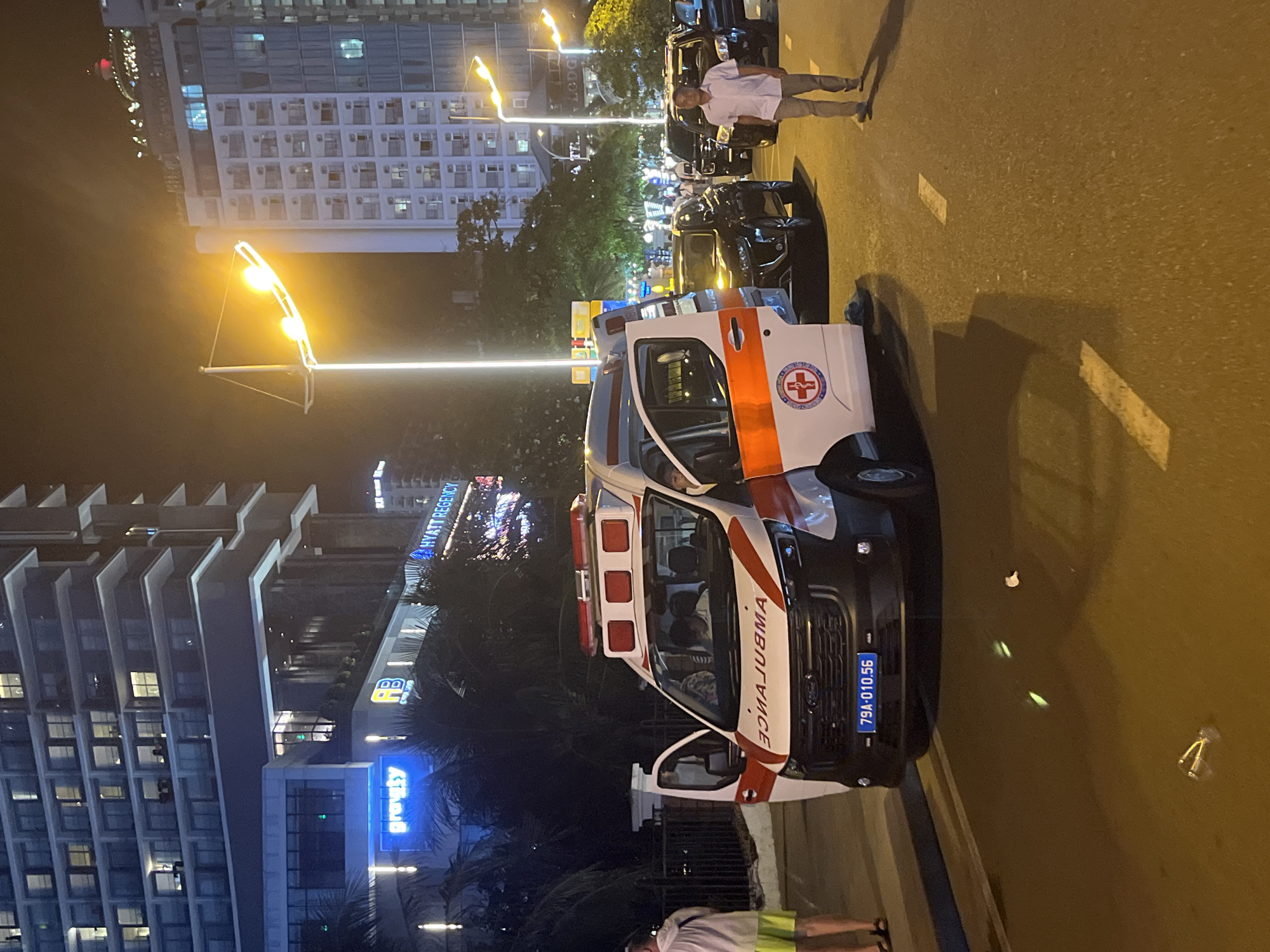 An ambulance is parked on a city street at night in Nha Trang, Vietnam. The ambulance, a white Ford Transit with red and orange markings, has its side door open, and a person is visible inside. A man in a light-colored shirt and pants walks away from the ambulance towards the right side of the frame. In the background, there are tall, modern buildings illuminated at night, including the Hyatt Regency hotel. Several other vehicles, including black SUVs, are also present on the street. The ground is paved, and streetlights cast a warm yellow glow on the scene. There are some debris on the road, including a plastic bottle and a crumpled piece of paper. The license plate on the ambulance reads "79A-010.56".