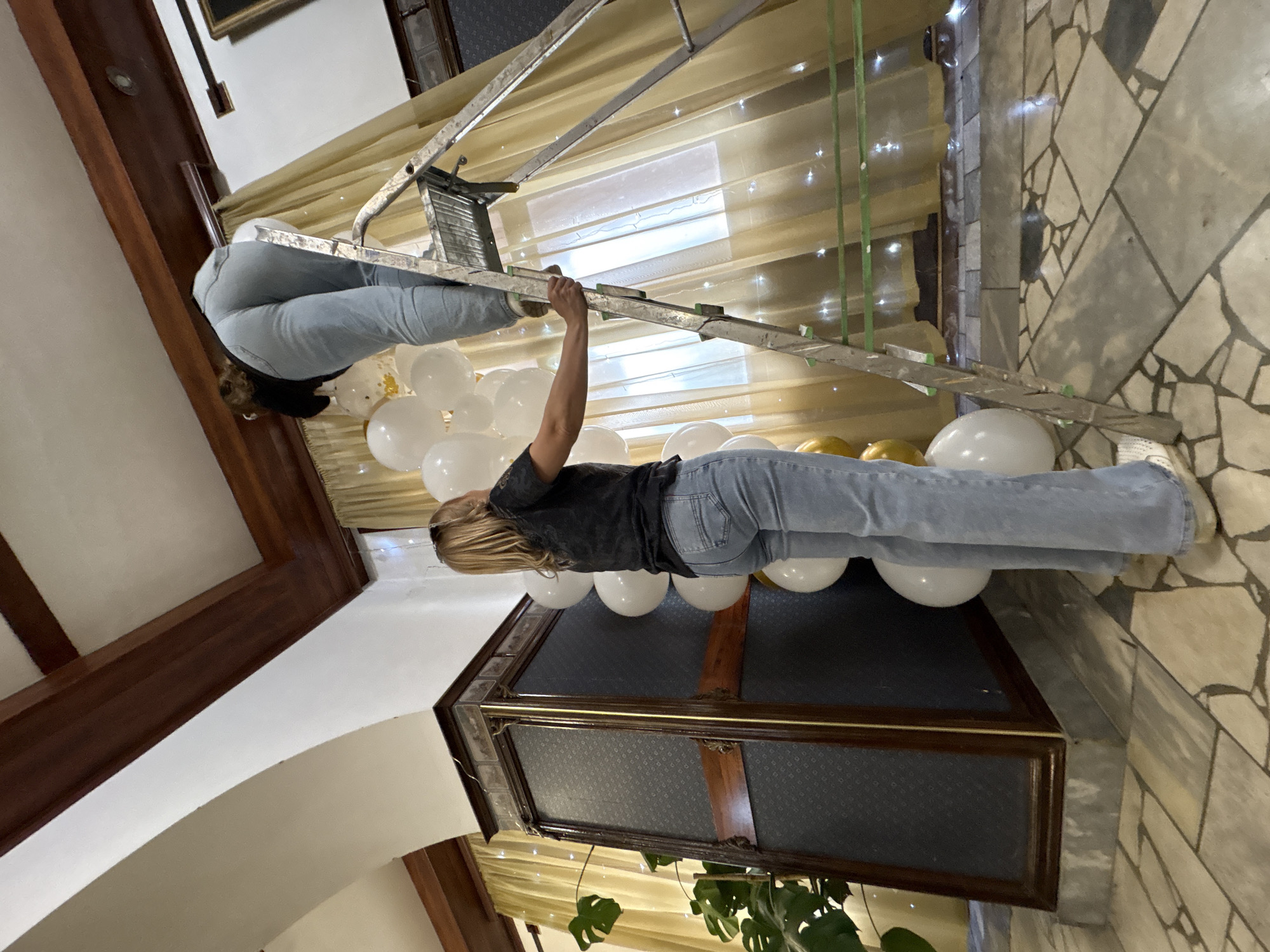 Two women are decorating with balloons in a room. The woman on the left, wearing a black t-shirt and light blue jeans, stands on the floor, holding a bunch of white balloons. The woman on the right, wearing a black top and light blue jeans, is on a silver ladder, reaching up to place a white balloon. The room has a tiled floor and cream-colored walls with dark wooden beams on the ceiling. Yellow curtains with string lights are visible in the background, and a large, ornate dark wooden cabinet is to the left of the frame. The scene is lit with artificial light. There is no specific indication of this scene being located in Zavod Mikhailovskii, Russia.