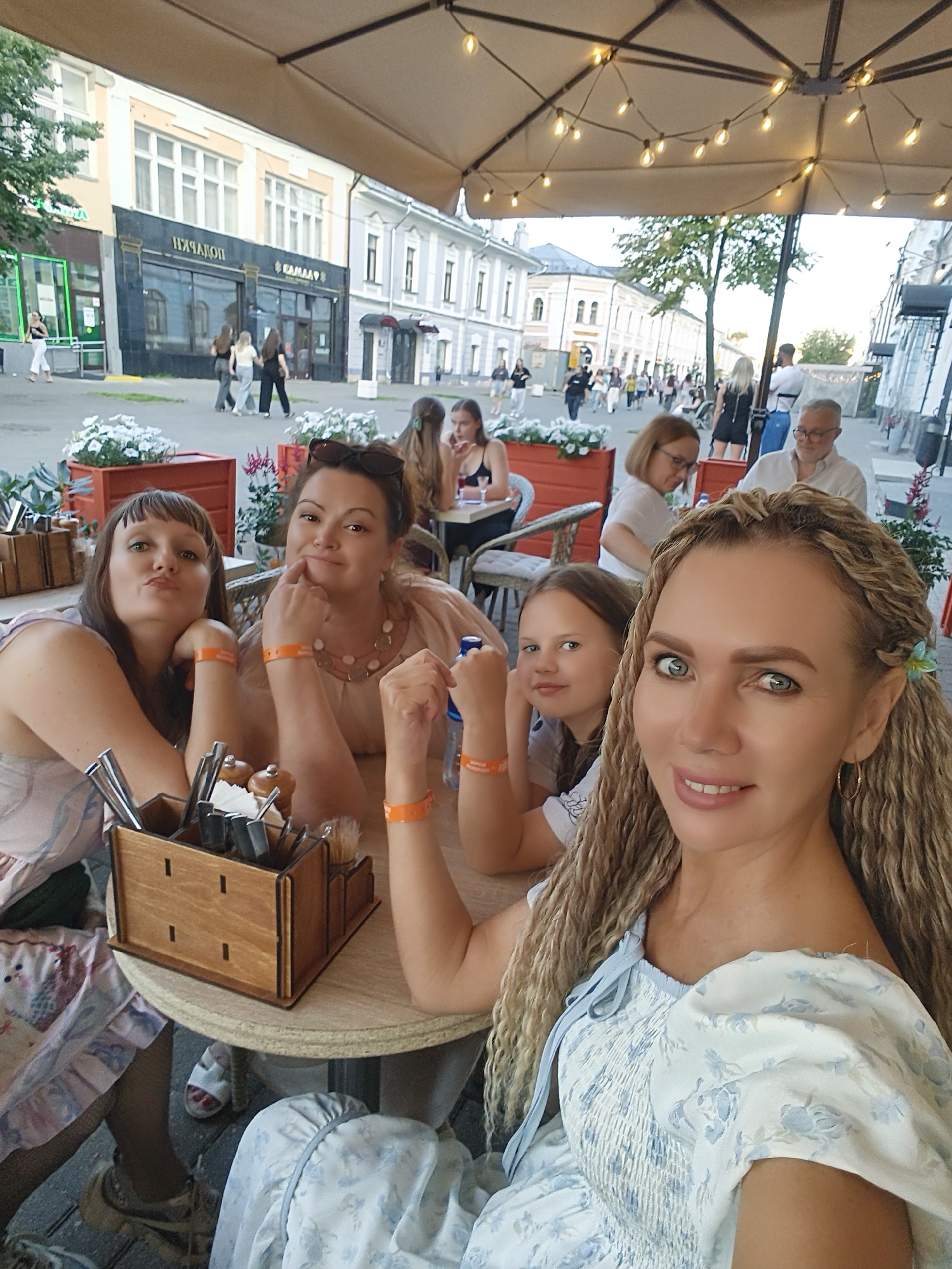 The image depicts an outdoor seating area of a cafe or restaurant on a street in Yaroslavl, Russia. A group of people, including women and a child, are seated at a table under a large umbrella adorned with string lights. In the background, a street lined with buildings is visible, with pedestrians walking along the sidewalk.
Specifically, the people in the foreground are a woman with blonde braided hair in a blue floral dress, smiling at the camera, and a young girl with brown hair. To their left are two other women. One has dark brown hair and is leaning on the table, looking towards the camera. The other, with light brown hair and glasses, is also seated at the table. Further back, a man with glasses and a white shirt is visible, and another woman with blonde hair is seated at a different table.
On the table, there is a wooden utensil holder filled with cutlery, salt and pepper shakers, and napkins. The environment suggests a pleasant outdoor dining experience, with potted plants decorating the area. The architecture of the buildings in the background indicates a historical urban setting.