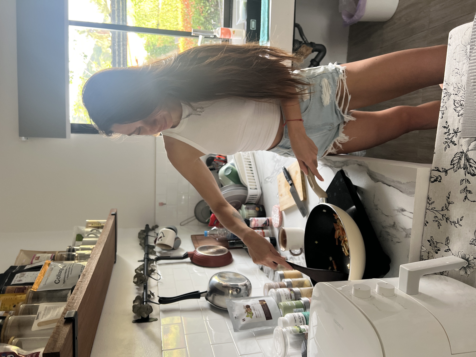 A woman stands in a kitchen in บ้านบางรักษ์, Thailand, cooking in a black pan. The kitchen has white tiled walls and countertop. On a shelf on the left are various jars and packages, likely containing spices and dry goods. Other cookware, including pots and pans, are visible on the countertop and hanging on the wall. A white air fryer is prominently placed on the right side of the countertop. Light streams through a window at the top of the frame, illuminating the kitchen. The woman is wearing a white tank top and denim shorts.