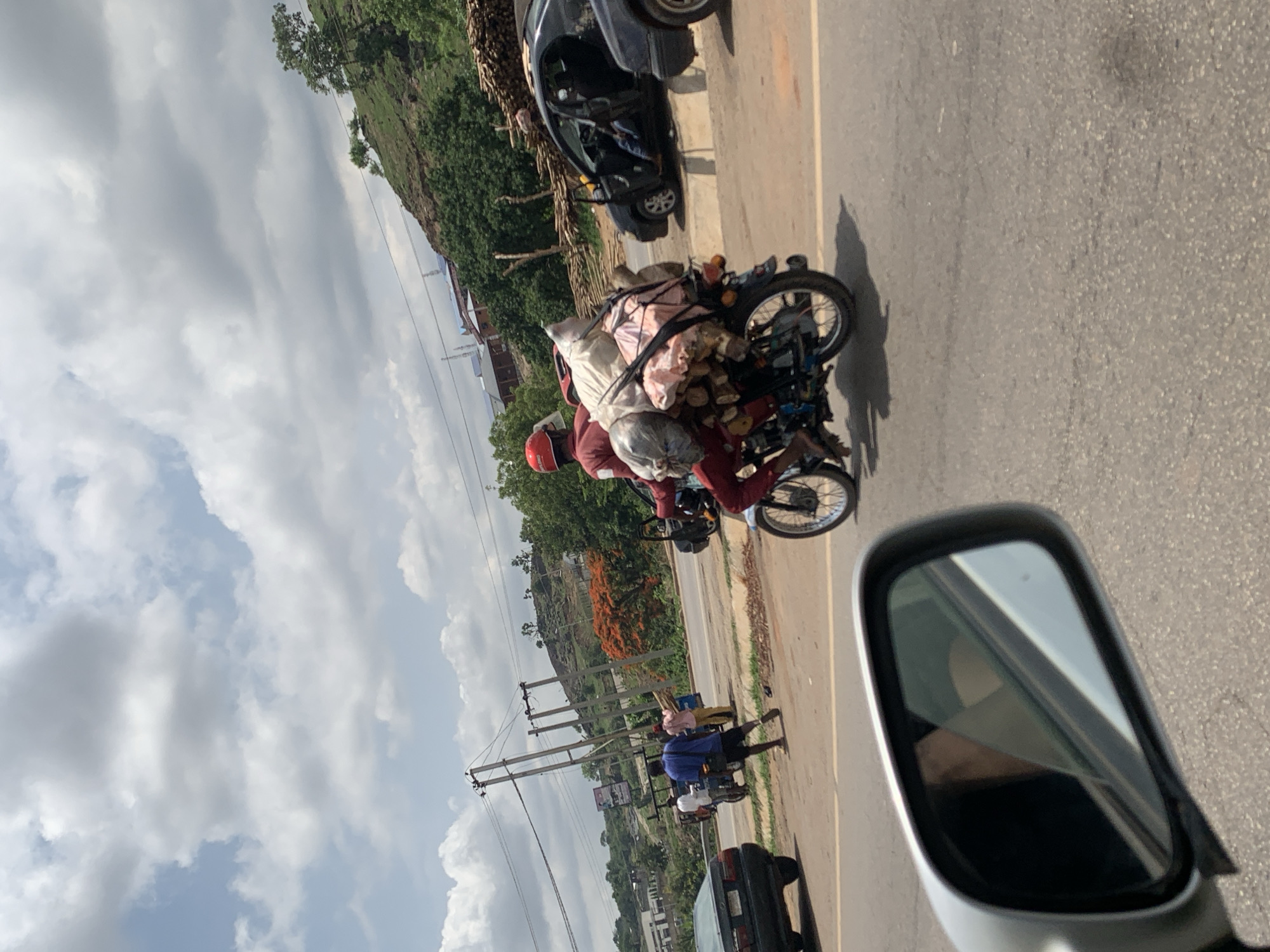 The image, apparently taken from inside a vehicle, shows a road scene in Nyanya, Nigeria. A motorcycle heavily loaded with bags and objects is moving on the road. The rider is wearing a red helmet and clothes. Other vehicles and pedestrians are visible along the road. Power lines are present along the road. The sky is cloudy. A side mirror of the car from which the picture was taken is visible.
