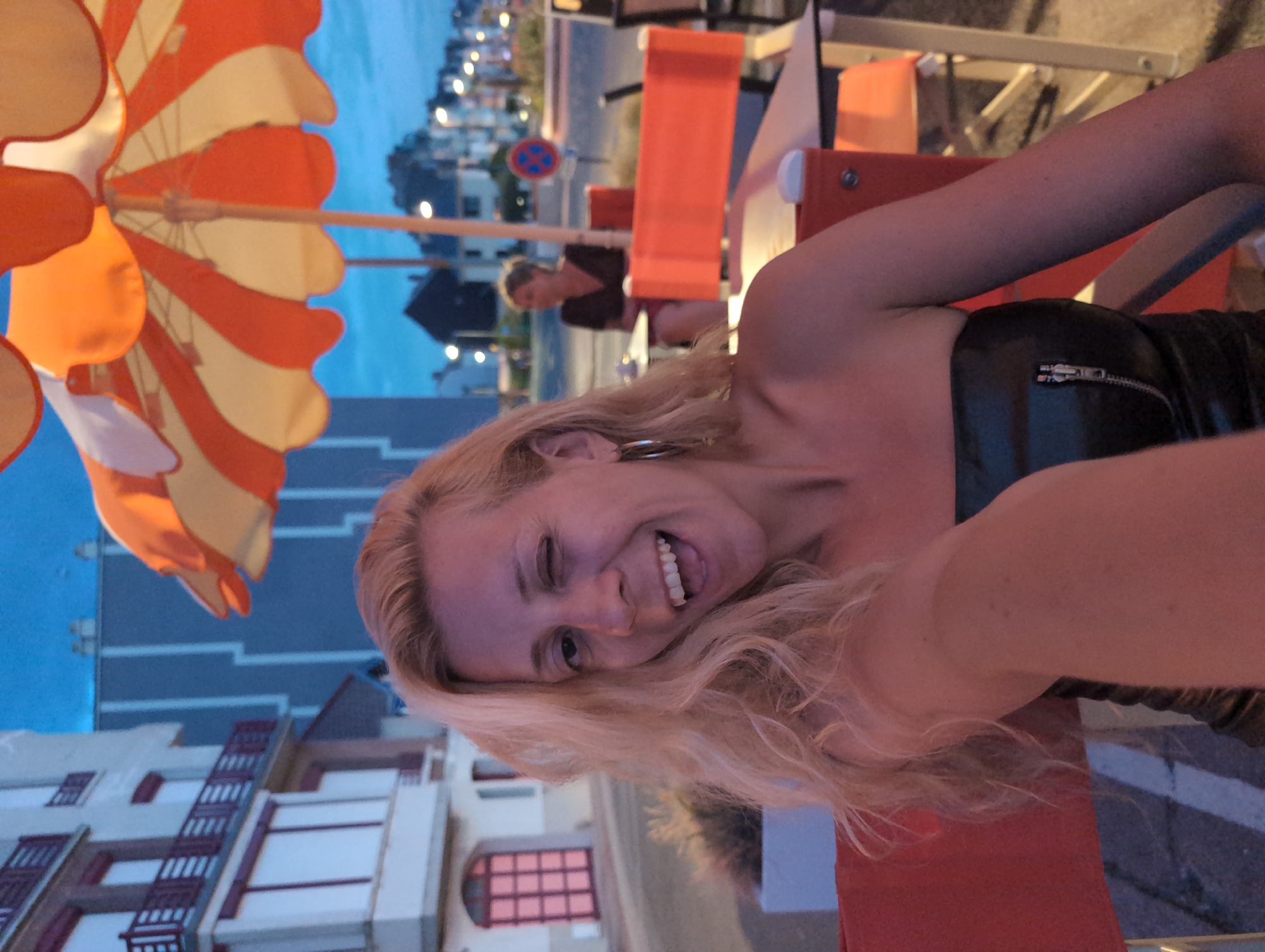 A woman with blonde, wavy hair is captured in a close-up selfie outdoors. She is winking with her right eye and smiling broadly, showing her teeth. She is wearing a strapless black top with a silver zipper detail. The background reveals a street scene at dusk in Mers-les-Bains, France, with a distinctive building featuring a light pink door and multiple windows. An orange and yellow striped umbrella is visible overhead, and further in the background, a woman is seated at a table. Streetlights and buildings line the street, suggesting a coastal town setting.