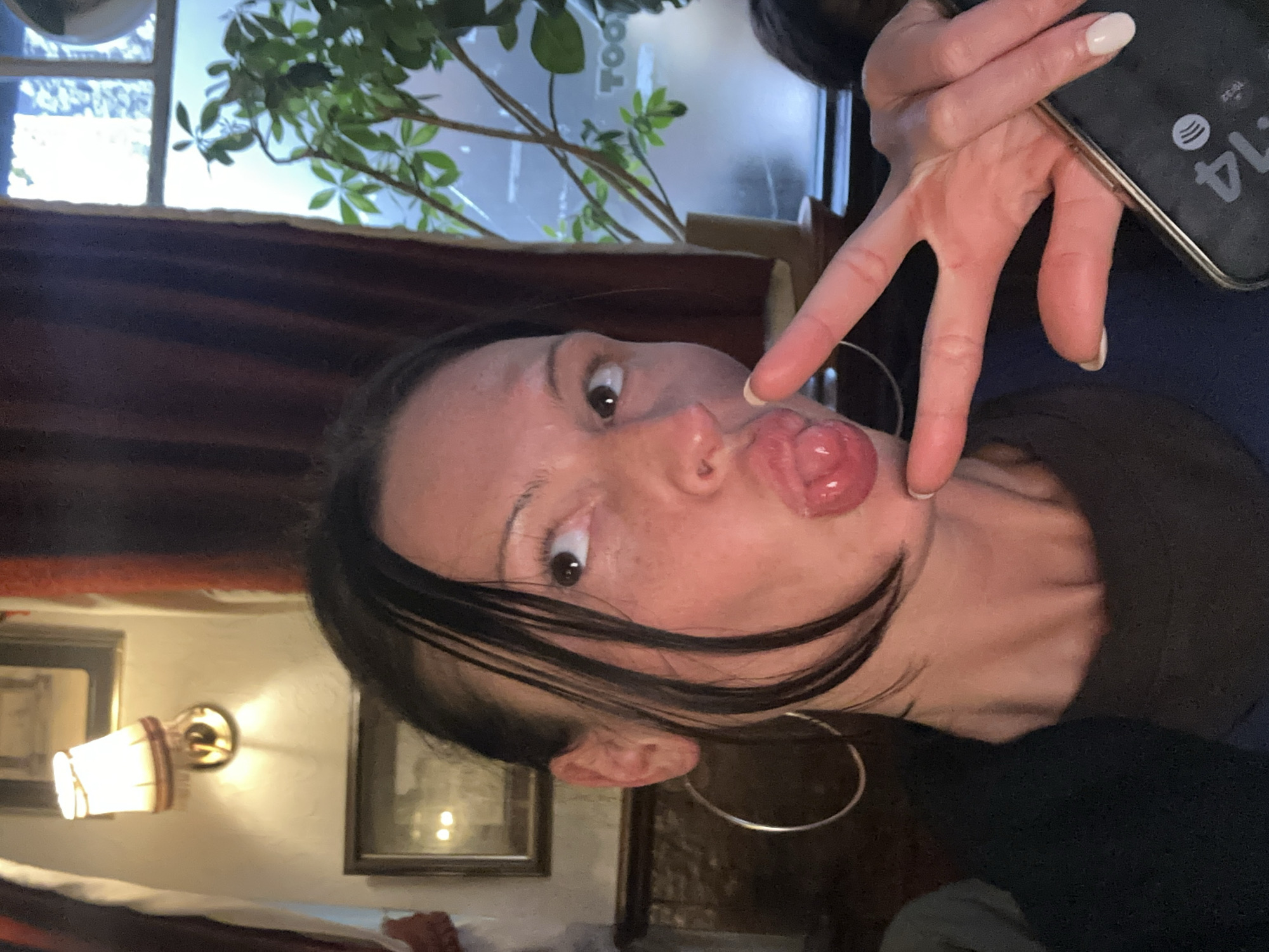 A medium indoor shot depicts a woman with dark hair and fair skin making a duck face and holding a smartphone with a time of 4:14 displayed, against a background featuring a window with a plant, a red curtain, a wall with framed art, and a wall lamp. The setting suggests a restaurant or pub, possibly in London, given the stylistic details and the type of establishment. The woman is likely taking a selfie.