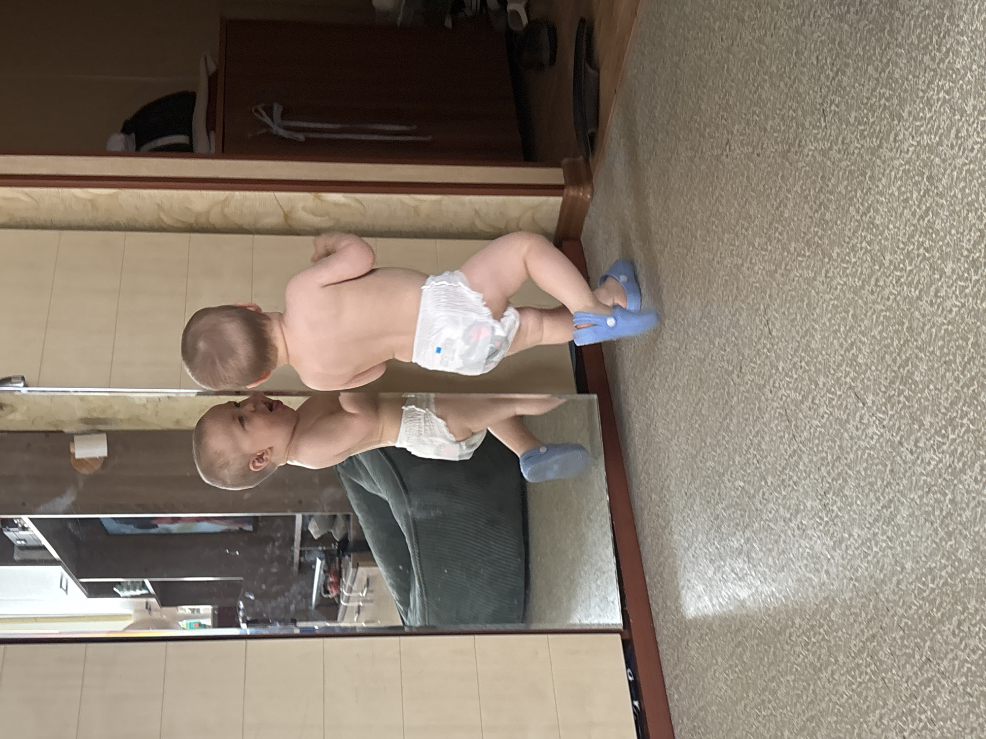 Two toddlers, each wearing a diaper and blue Crocs, are in a room with a large mirror. The toddler on the left is facing the mirror and appears to be crying or making a distressed face. The toddler on the right is facing away from the camera and towards the mirror, with their back to the viewer. They are wearing a diaper and blue Crocs, and their arms are raised slightly.
The room has a light-colored carpeted floor and a beige, textured wallpaper on the right side of the frame. To the left of the mirror, a tall, light-colored wardrobe is visible. In the reflection of the mirror, a large, dark green beanbag chair is prominent, as is a shelf unit with various items. The background of the reflection suggests an indoor environment, possibly a living room or bedroom.
The image does not provide any information that would indicate the location within the city of Naberezhnye Chelny, Russia.