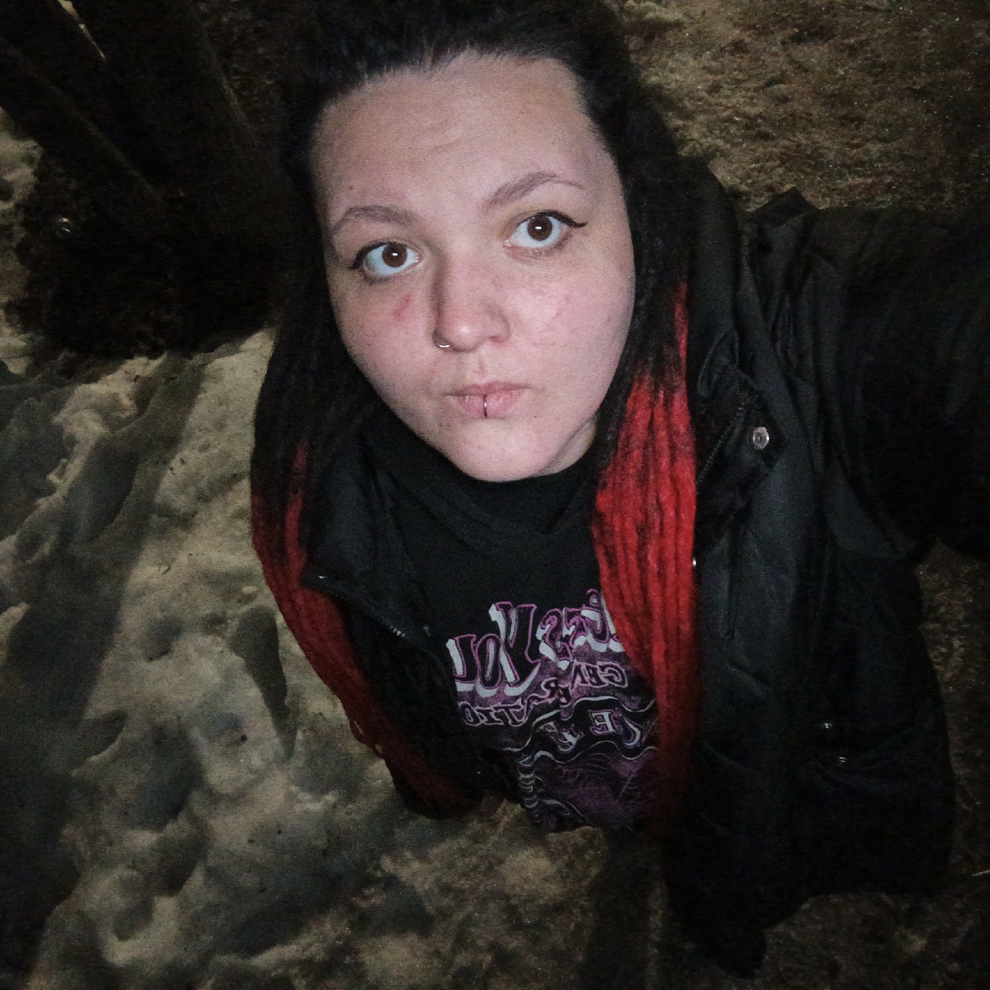 A selfie photograph, taken from a low angle, depicts a fair-skinned individual with dark hair streaked with red. The person has a nose ring and a labret piercing. Their eyes are brown and lined with eyeliner. There is some redness on their face, particularly around the left eye. They are wearing a black t-shirt with a purple and white graphic design under a black, puffy zip-up jacket. The setting appears to be outdoors, with snow and some dark objects, possibly tree trunks, visible in the background. Although the background is blurry, this image was taken in Chaikovsky, Russia.