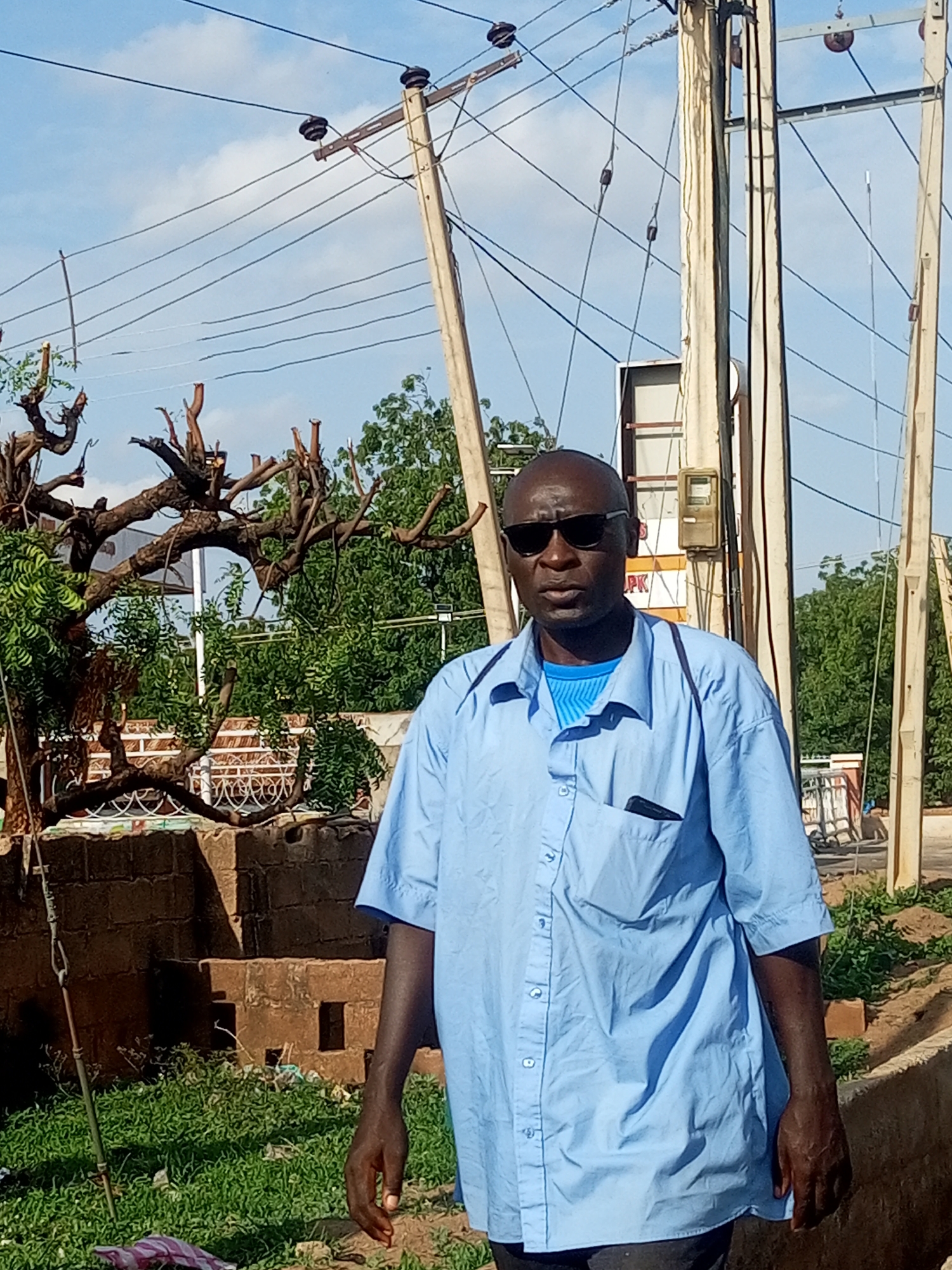 A man stands outdoors in front of utility poles and wires. He is bald, wears dark sunglasses, and a light blue short-sleeved button-down shirt over a blue t-shirt. A black pen is tucked into the chest pocket of his shirt. He has a dark complexion and is looking towards the camera with a neutral expression.
The background shows several wooden utility poles with numerous wires strung between them. Some of the poles appear weathered and aged, with evidence of sun damage. Behind the poles, there are trees with green foliage, some of which are bare and pruned. In the mid-ground, there's a structure made of brick or stone, and further back, a glimpse of a building with a white panel. The ground in front of the man is covered in green grass and some scattered debris. To the right of the man, a concrete edge is visible.
The lighting suggests it is daytime, likely with clear or partly cloudy skies, as indicated by the bright sunlight and shadows. The presence of utility poles and the style of the buildings hint at a location in Nigeria, consistent with Gombe. No specific activity is taking place; the man appears to be posing for the photo. There is some text visible on a white sign behind the man, partially obscured by the utility pole, with the letters "PK" clearly legible.
