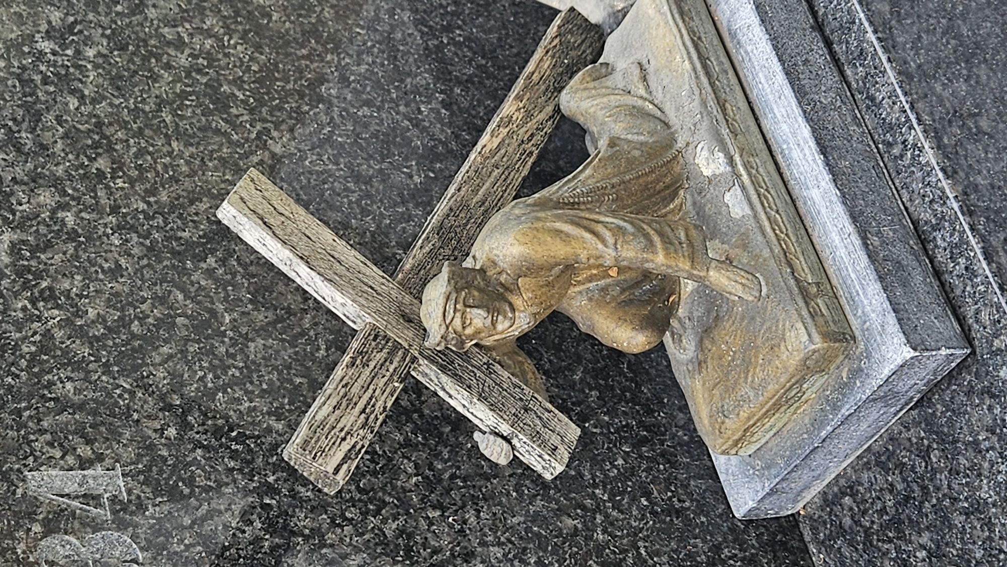 The image shows a close-up, eye-level shot of a statue depicting Jesus Christ carrying the cross. The statue is made of a bronze-like material and is weathered, with some patina and wear visible. Jesus is shown kneeling, with his right knee on the ground and his left leg bent, supporting the weight of the cross. The cross itself is depicted as made of rough, aged wood. The statue is placed on a rectangular base, which is resting on a larger, rectangular stone slab. The background is a dark, speckled granite surface, typical of gravestones, with the numbers "34" faintly visible etched into it on the left side. The scene appears to be located in a cemetery, possibly the Catholic Cemetery in Prešov, Slovakia, given the style of the monument. The lighting is natural, casting subtle shadows that highlight the textures of the materials.