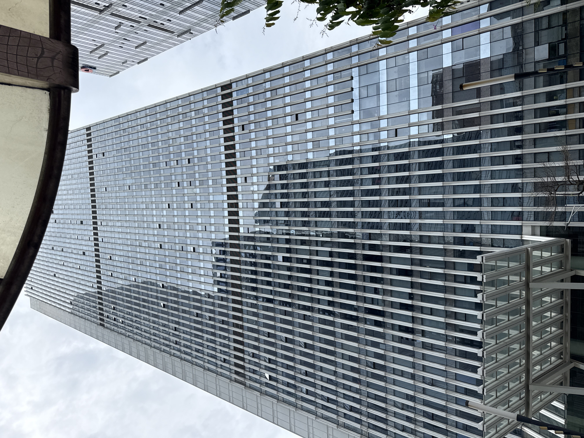 A low-angle, full-frame shot captures the exterior of a modern, glass-facade skyscraper in Tianhe District, China. The building's facade is composed of numerous vertical metallic fins interspersed with rectangular glass panels. Some of the glass panels appear to be open. The skyscraper is tall and slender, with its peak extending out of frame. Reflections of other buildings and the overcast sky are visible on the glass surfaces. A second, partially visible skyscraper with a similar glass facade stands to the right of the main building. In the foreground, a section of a glass and metal canopy structure is visible at the bottom of the frame. The sky is a uniform gray, indicating an overcast day. No people are visible in the image.