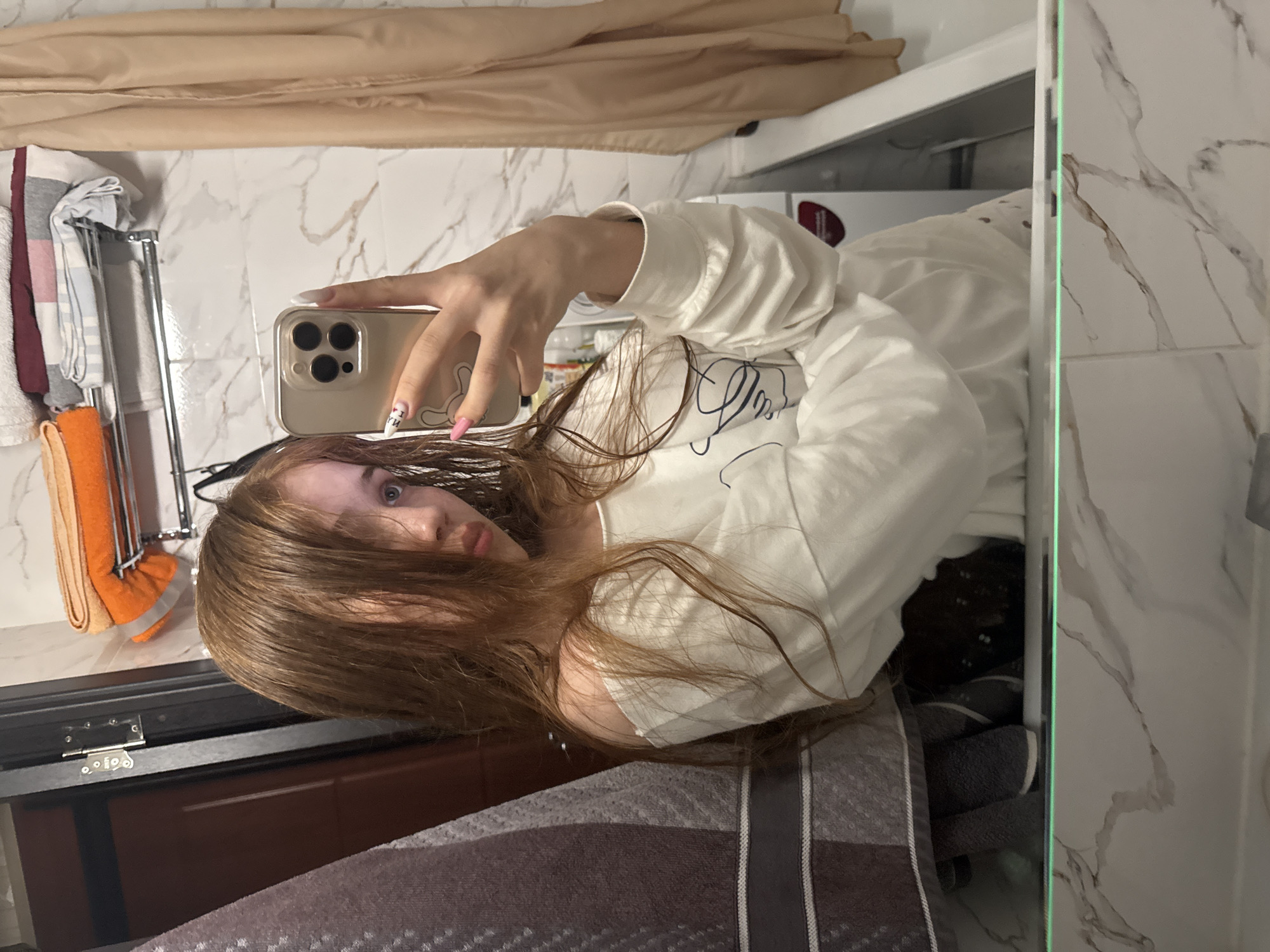The image displays a mirror selfie taken by a single individual in what appears to be a bathroom. The person, a young individual with long, reddish-brown hair that is partially wet or styled forward to obscure a portion of their face, is wearing a long-sleeved white top, possibly a sweatshirt, which is pulled down on the left shoulder to expose it. A dark, abstract line drawing is visible on the front of the shirt. The individual holds a gold-colored smartphone, likely an iPhone Pro model due to its triple camera array, in their right hand, angled to capture their reflection. The phone is encased in a clear cover featuring a white bunny or similar cartoon character on the back. Visible fingernails are manicured; the thumb displays a pink base with a white design, while the index finger has a white base with a red and black design.
The background, reflected in the mirror, shows white marble-effect wall tiles with grey-brown veining. To the viewer's left, a dark grey and white striped towel or fabric hangs. A dark brown door frame with silver hardware (hinge and latch mechanism) is visible. Above the individual's head, a chrome or silver metal shelf holds several folded towels in various colors, including orange, white, red, and blue-striped. To the viewer's right, a light beige or tan fabric, possibly a shower curtain, hangs alongside a white porcelain fixture, likely a bathtub or sink. A white household appliance, potentially a washing machine, is partially visible further to the right. The lower edge of the mirror shows a horizontal green light strip. The image does not contain any visual markers or identifiable features to confirm the location as Naberezhnye Chelny, Russia.