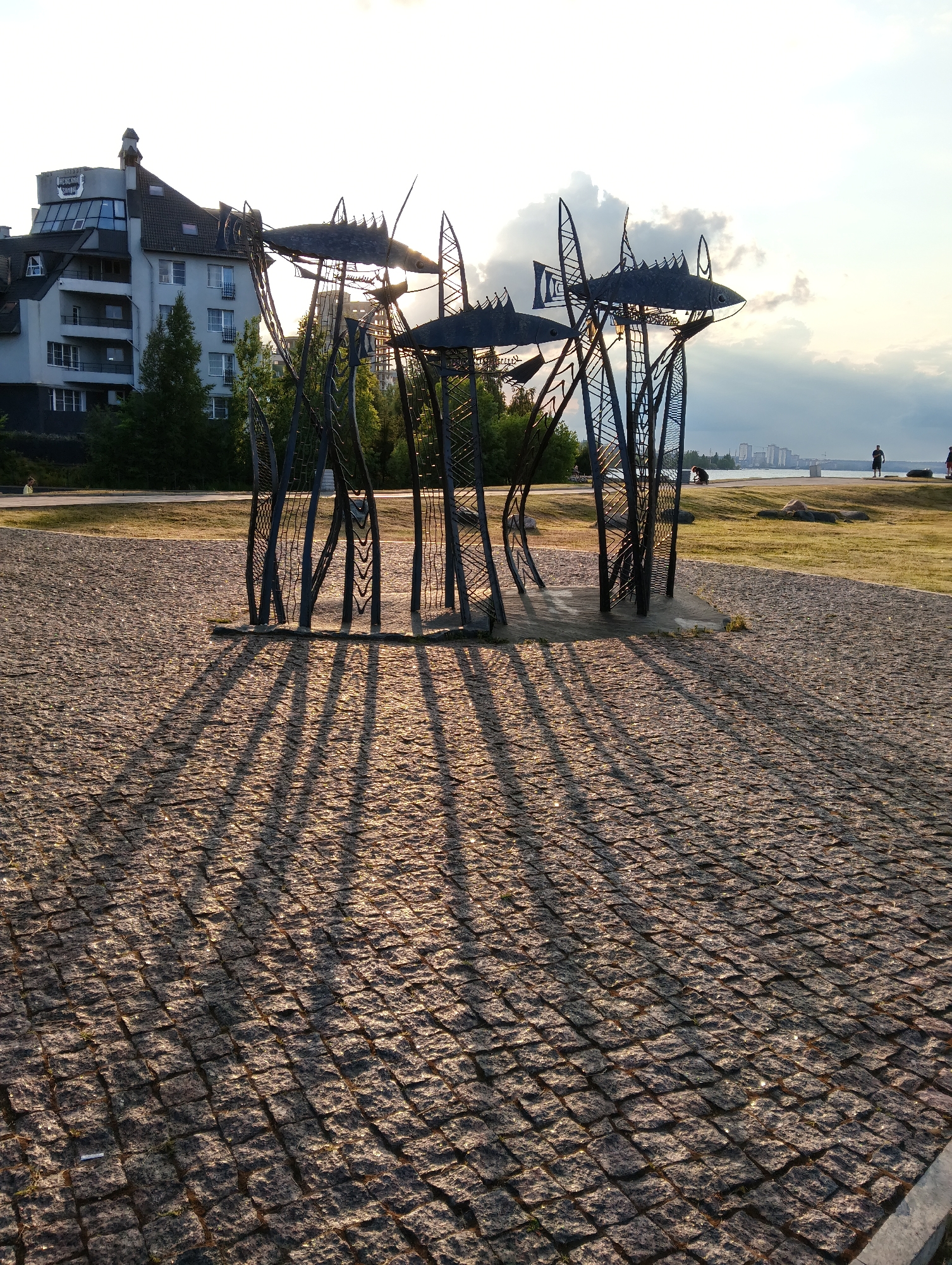 The image shows a public sculpture made of black metal, resembling stylized fish on tall stalks. The sculpture is situated on a cobblestone plaza. In the background, there is a modern, multi-story building with a sign that appears to read "ОНЕЖСКАЯ РЫБА" (Onezhskaya Ryba), suggesting the location is near Lake Onega in Petrozavodsk, Russia. A tree-lined embankment and the calm waters of a lake or bay are visible beyond the plaza. The sun is low in the sky, casting long shadows from the sculpture onto the cobblestones, indicating it is late afternoon or early evening. A few people are visible in the distance walking along the waterfront.