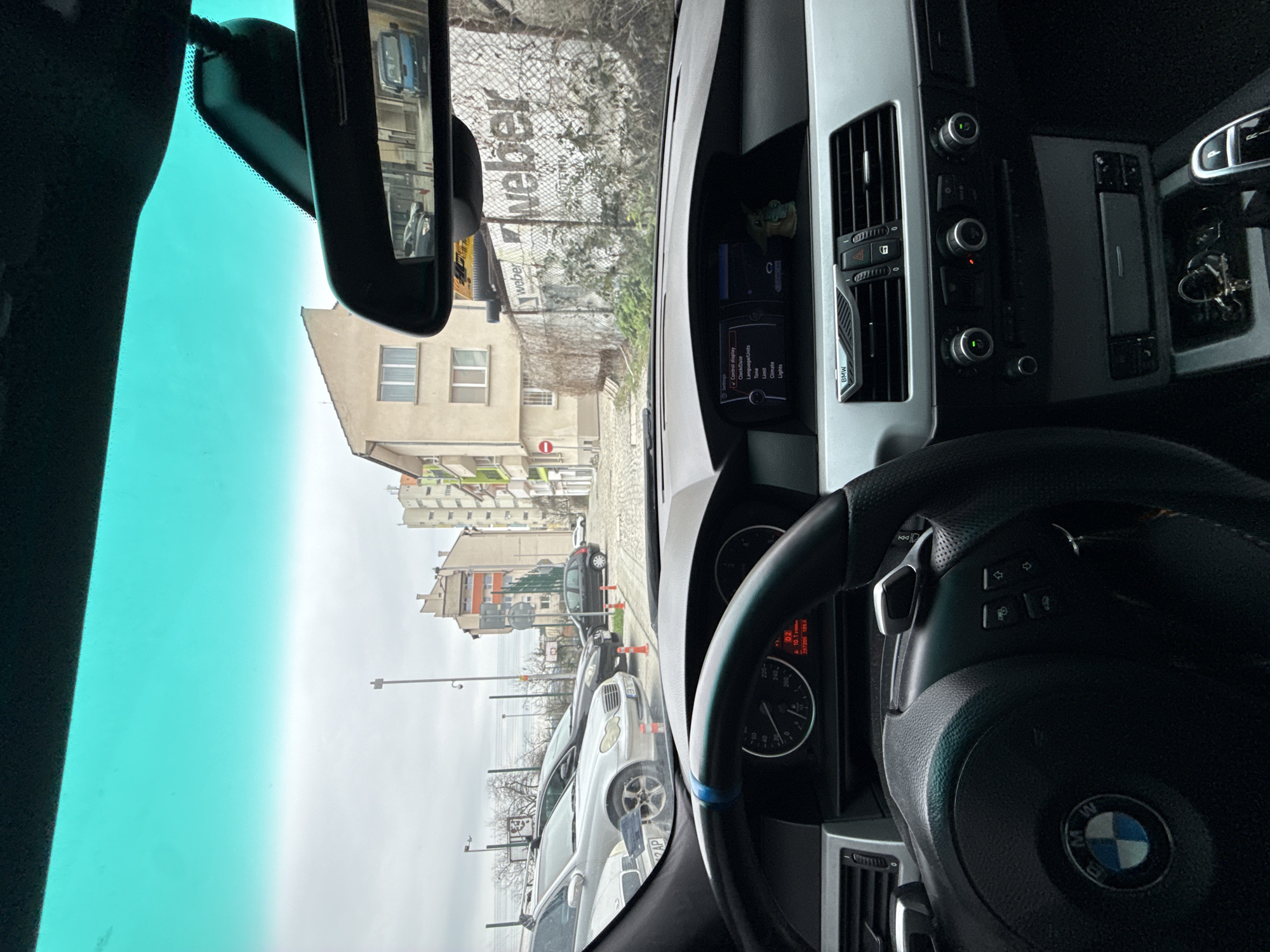 The image shows a view from inside a car, likely a BMW, looking out onto a street in what appears to be an urban environment. The car is parked, with several other vehicles visible in front. There is a building in the background. A sign with the word "weber" is also visible. The dashboard and steering wheel of the car are prominent in the foreground. The navigation display says 'Clock/Date', 'Language/Units', 'Limit', 'Climate', 'Lights'. Based on the license plate 'D' and other details, the location is likely Gabrovo, Bulgaria.