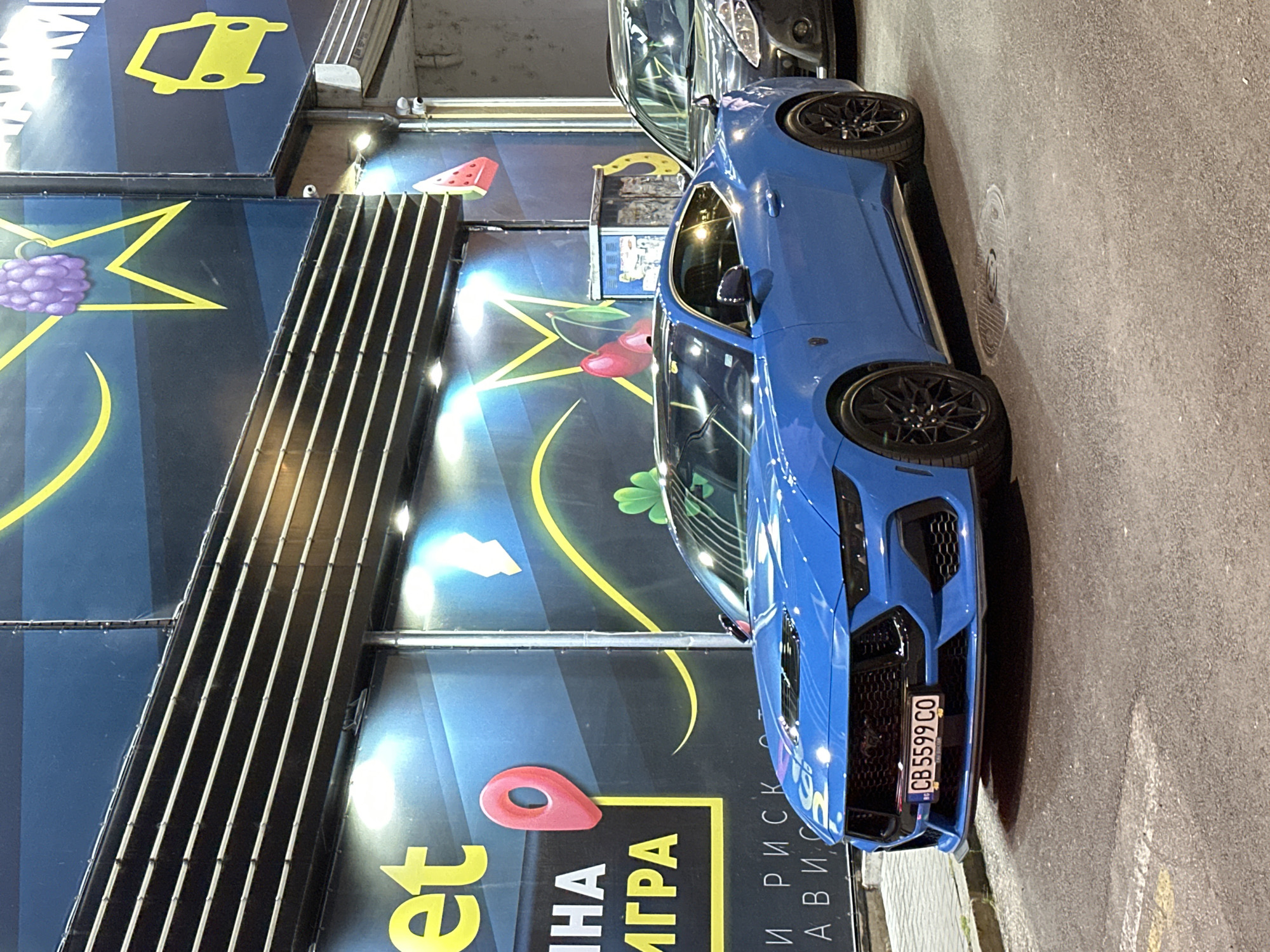 A blue sports car is parked on a street next to a building with large advertisements. The car is positioned lengthwise along the right side of the frame, facing away from the viewer. It has black rims and a black grill. A license plate on the front reads "CB 5599 CO". The building on the left has a dark blue background with various graphic elements including a yellow lightning bolt, a purple bunch of grapes, and a red pin icon. There are also strips of vertical lights on the building's facade. Text is visible on the building, with some letters in yellow and white. Part of the text includes "et", "И НА", "ИГРА", and "И РИСК АВИС". The scene appears to be at night or in low light, as the building's lights are illuminated. The car is parked on a paved road with some painted white lines. The overall impression is of a street scene in an urban environment.