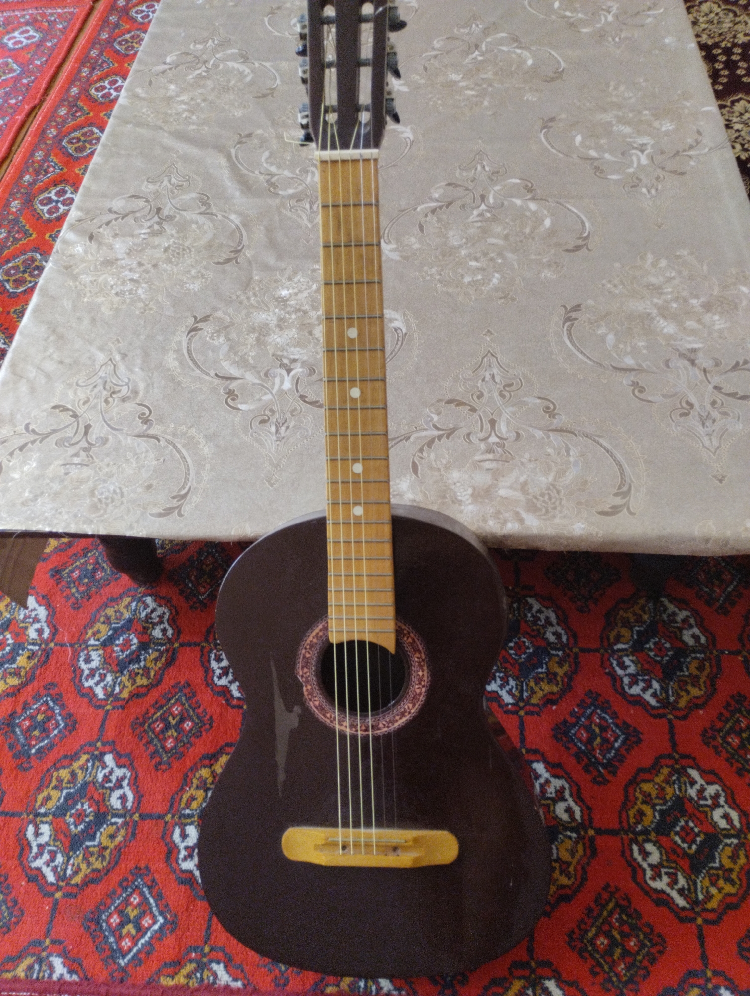 A dark brown acoustic guitar rests vertically on a red carpet with intricate geometric patterns. The guitar is positioned on a table covered with a light-colored tablecloth featuring a damask pattern. The scene appears to be indoors, possibly a home, given the presence of the carpet and table. The guitar's neck is at the top of the frame, with its headstock and tuning pegs visible. The body of the guitar, including the soundhole and bridge, is towards the bottom. The overall impression is a still life composition, with the guitar as the central subject. The location of Yangibazar, Uzbekistan, is not visually identifiable from the image itself.