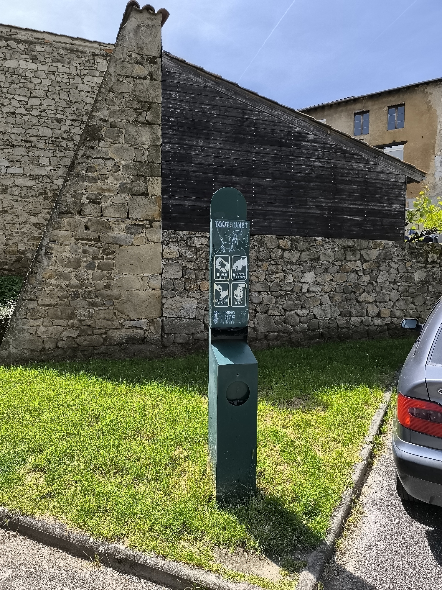 The image shows a "Toutounet" pet waste disposal station in Pélussin, France. The station is green and features instructions for using a bag to pick up and dispose of pet waste. It stands on a small patch of green grass bordered by pavement. Behind the station are a stone wall and a dark, wooden-clad structure, potentially a building facade. A gray car is partially visible to the right of the station. The sky is blue with faint contrails.