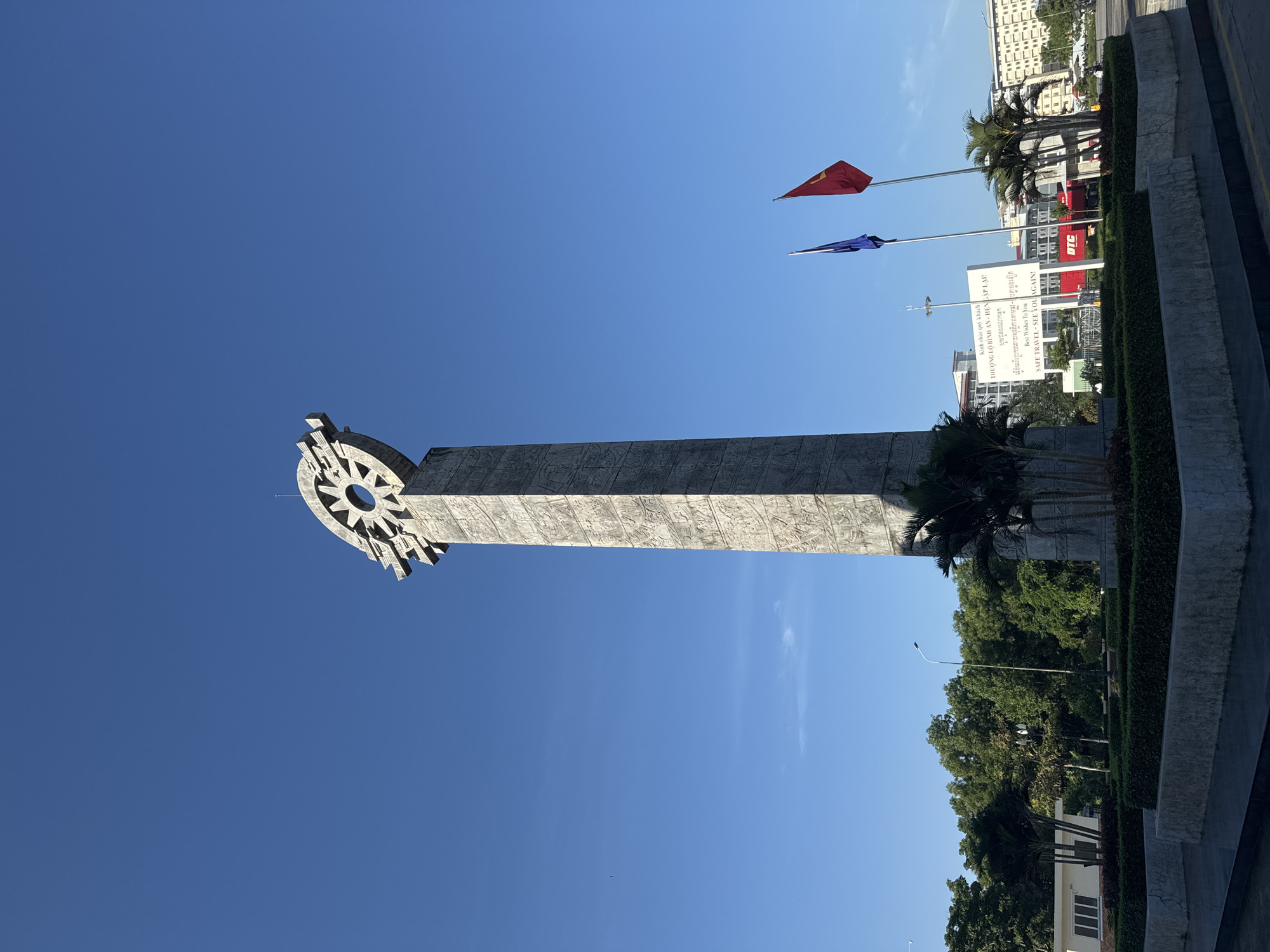A tall, concrete monument with intricate carvings stands against a clear blue sky in the city of Ben Cau, Vietnam. The monument is topped with a decorative circular structure. In front of the monument, there are well-maintained hedges and small palm trees. Two flagpoles stand nearby, one with a blue flag and the other with a red flag, the latter appearing to be the Vietnamese flag. Behind the monument, a white sign displays text in both Vietnamese and English, with the English portion reading "Best Wishes To You SAFE TRAVEL - SEE YOU AGAIN!". A red shipping container with the logo "OTC" is visible behind the sign. The background shows buildings, trees, and a street with some traffic, suggesting an urban environment. The sun is shining, casting shadows.