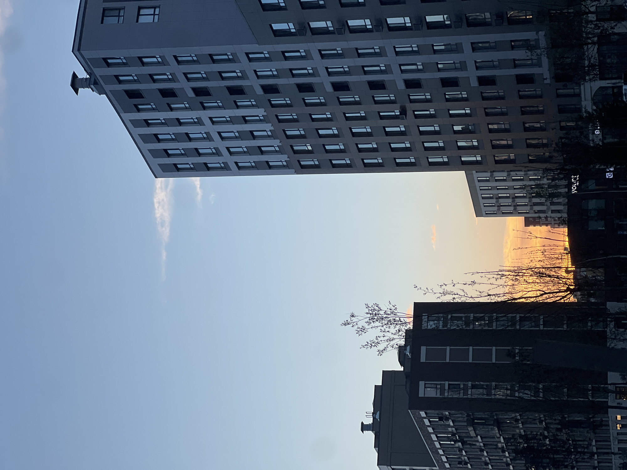 The image captures a modern urban scene at dusk in Astana, Kazakhstan, likely in a newly developed residential area. The foreground features the stark silhouettes of several multi-story apartment buildings against a gradient sky transitioning from a pale blue at the top to warm oranges and yellows near the horizon. The buildings are characterized by their contemporary architecture, featuring numerous windows and clean, geometric lines. The tallest building on the right is a light grey structure with a repeating pattern of dark-framed windows. To the left, a darker, taller building is visible, also with many windows. Further to the left and slightly in the background, a shorter, dark-colored building with brick accents is present. Bare trees, with delicate branches reaching upwards, are sparsely scattered in front of the buildings, their forms silhouetted against the twilight sky. A faint wisp of cloud is visible in the upper left portion of the sky. The overall impression is one of urban development and the quiet transition from day to night.