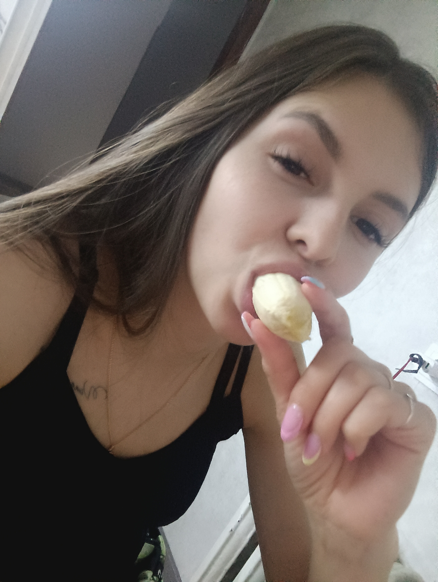A young woman is eating a peeled banana. She is wearing a black tank top and a delicate gold necklace. She has long, wavy brown hair and manicured nails with pink and yellow accents. The background is blurry, but appears to be an indoor setting with light-colored walls. The location is unknown.