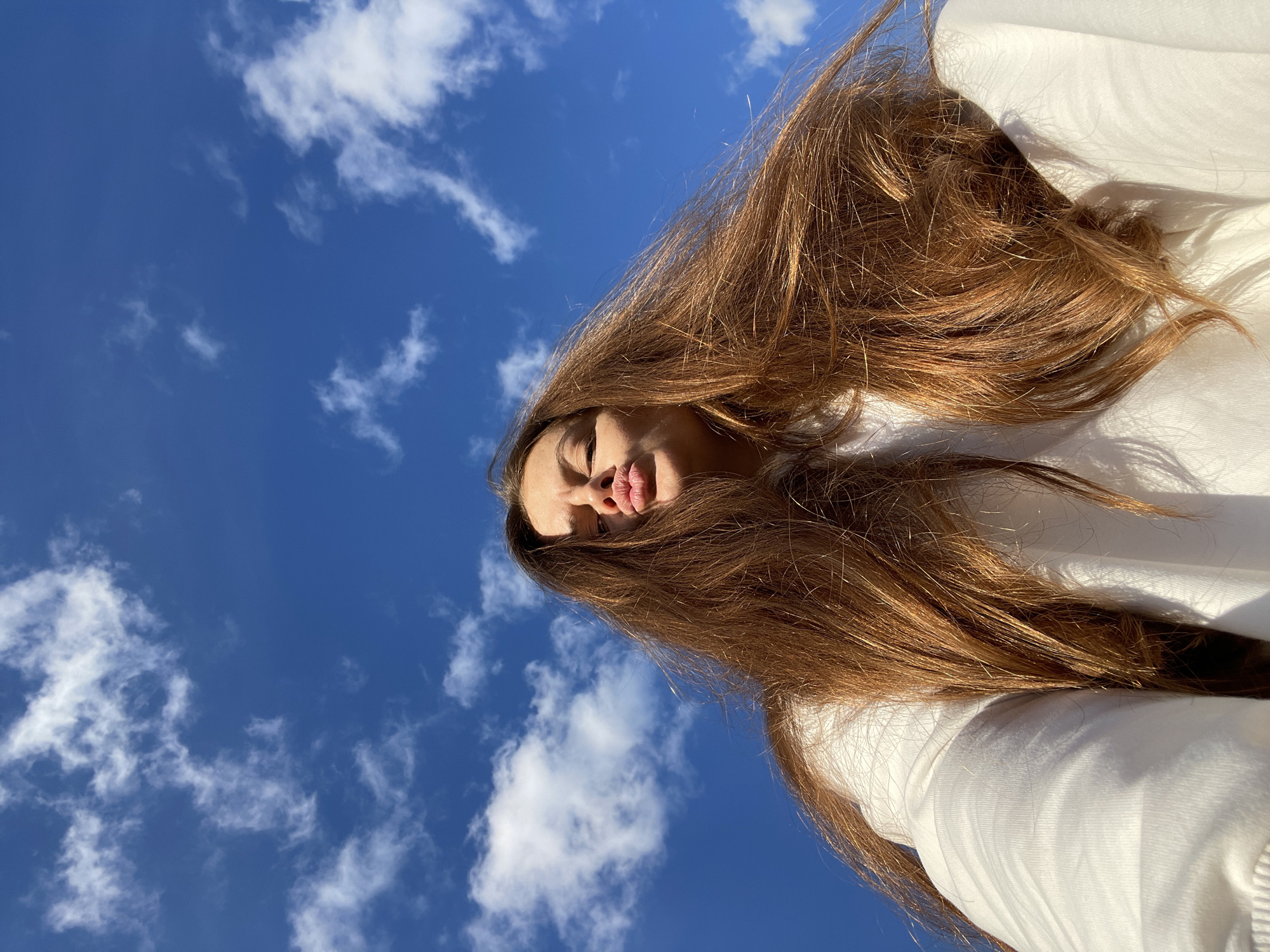 The image is a low-angle, close-up shot of a young woman, likely taken outdoors in Заречный, Russia. She has long, reddish-brown hair that cascades around her face and across a white garment, possibly a shirt or sweater. The woman is puckering her lips, as if blowing a kiss or making a playful face. Her eyes are partially closed, and the sunlight casts a shadow across her forehead.
The background is a vibrant, bright blue sky dotted with fluffy white clouds. The lighting suggests it is daytime, likely late morning or early afternoon, given the intensity of the sunlight and the clear sky. The overall mood of the image is cheerful and spontaneous, capturing a moment of lightheartedness. There is no visible text in the image. The composition, with the subject's face partially obscured by her hair and the extreme low angle, creates an intimate and engaging perspective.