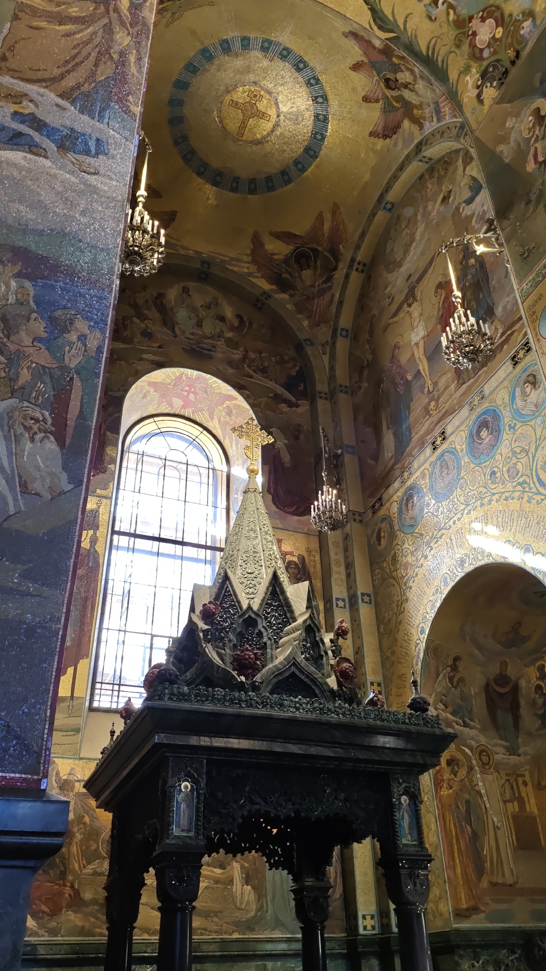 This media file shows the interior of a grand, ornately decorated church, likely in Saint Petersburg, Russia. The scene is dominated by rich mosaics and frescoes covering the walls and ceiling, depicting religious figures, angels, and scenes. A large, arched window with a grid pattern allows natural light to illuminate the space, suggesting it is daytime.
In the foreground, a dark, intricately carved wooden structure stands on thick pillars. This structure appears to be a monument or altar, topped with a decorative, tiered pyramidal element adorned with red flowers and topped with a golden cross. The detailed carvings on the structure suggest significant craftsmanship.
Hanging from the high, vaulted ceiling are two ornate chandeliers, casting warm light and adding to the opulent atmosphere. The ceiling itself is a canvas of golden mosaics, featuring a central circular mosaic with a cross and further decorative motifs. The walls are covered with a continuous narrative of painted scenes and mosaics, adding depth and color to the environment. The overall impression is one of immense historical and artistic significance, with a solemn yet awe-inspiring atmosphere.