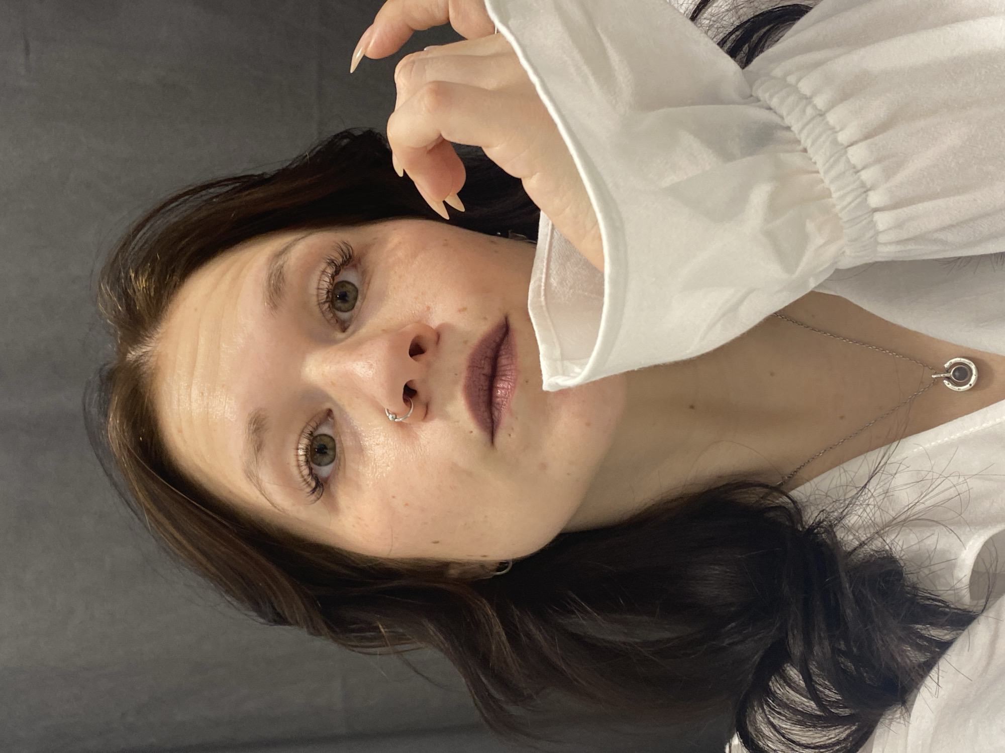 A young woman with long dark hair, light skin, and green eyes, is positioned in the center of the frame. She is wearing a white blouse with long sleeves and a silver necklace. She has a silver nose ring. Her left hand is near her face, with her fingers slightly curved. The background is a plain grey. The location in the city of Chaikovsky, Russia, is not verifiable from the image alone.