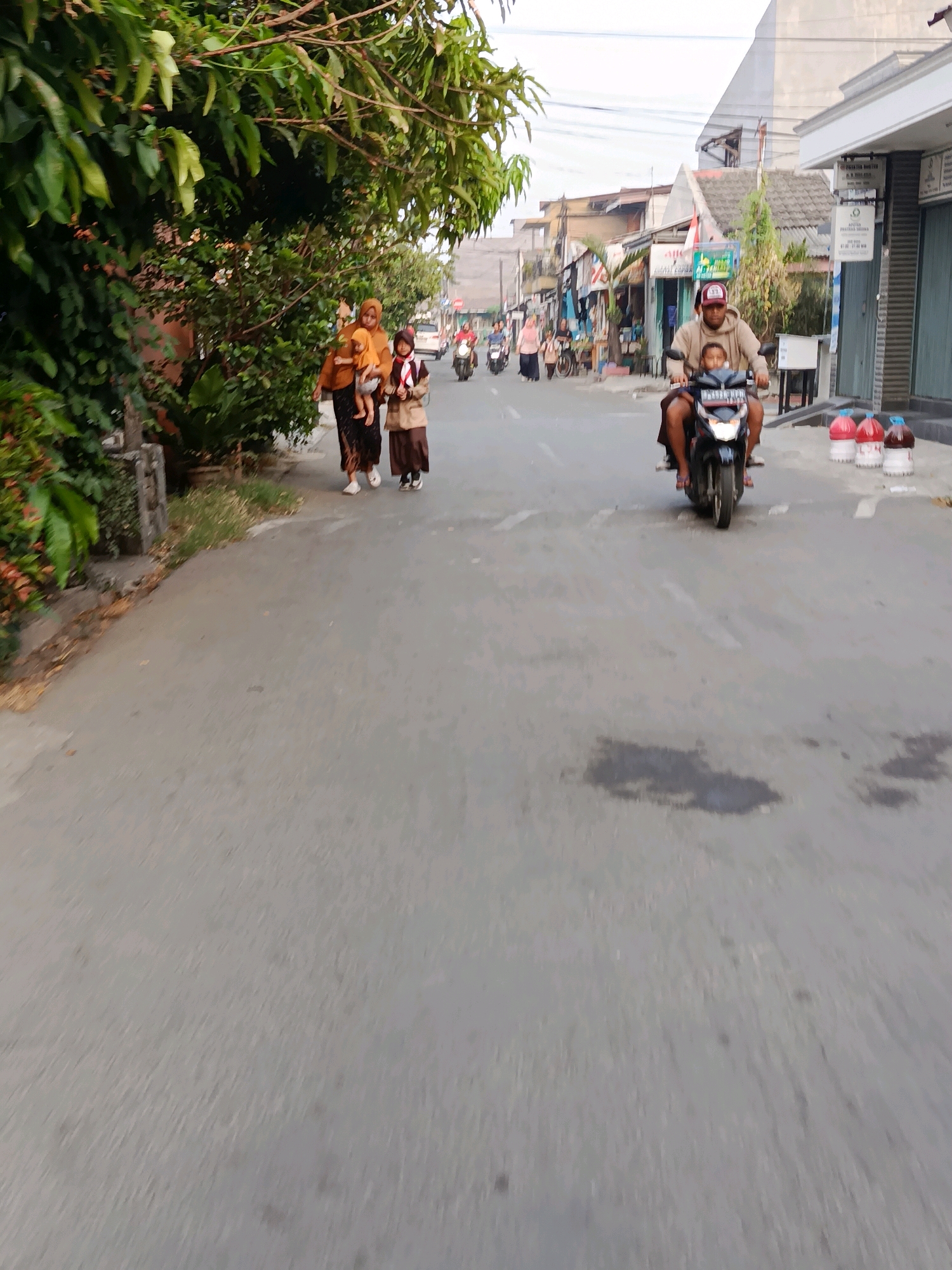 In Bekasi, Indonesia, a family walks along a paved road. A woman in a hijab and a long brown dress carries a baby. Next to her, a young girl in a school uniform walks along. They are walking on the side of the road, adjacent to a dense line of trees. In the distance, a man and a child ride a motorcycle. The man is wearing a beige hooded sweatshirt and a baseball cap, while the child is seated in front of him. Several other motorcycles are also visible on the road, as well as buildings on the right side of the street. The sky is overcast. Three large plastic bottles containing red and white liquid are placed on the side of the road near the buildings.