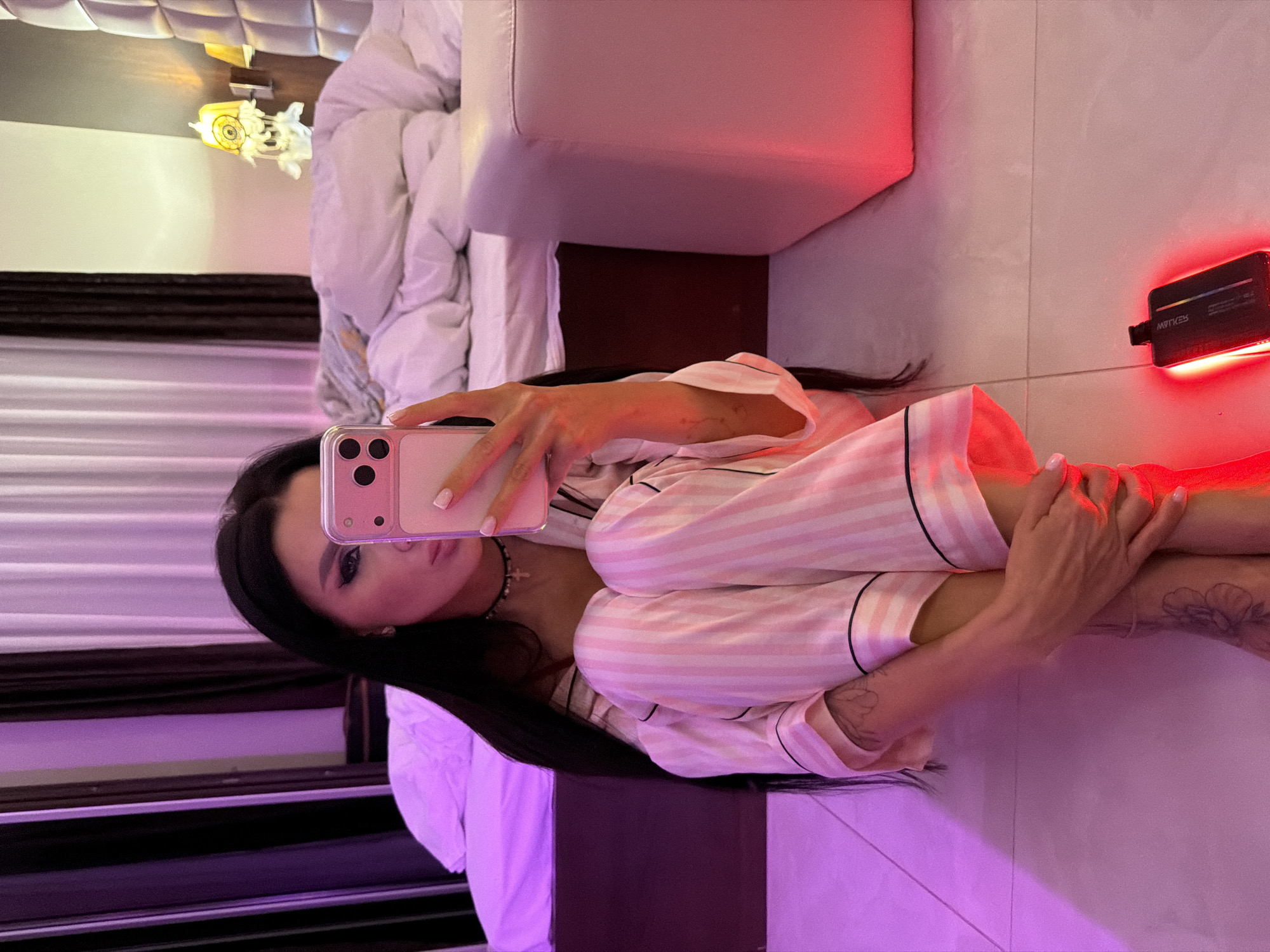 A woman is captured in a selfie taken in a dimly lit indoor setting, likely a bedroom or hotel room in Boreti, Montenegro. She is seated on a light-tiled floor, with her legs drawn up towards her chest, and holds a light pink smartphone with a matching case, seemingly taking her own picture.
The subject has long, dark hair and is wearing pink and white striped pajamas with black piping. Her left eye, visible in the reflection, features dark eye makeup. Her hands display manicured nails with white tips. Tattoos, appearing to be floral patterns, are visible on her forearms. She also wears a dark choker necklace adorned with a small cross pendant.
The background includes a bed with a padded headboard and white bedding, next to which sits a light-colored armchair or sofa. A small, decorative yellow wall lamp is visible near the bed. The room's ambiance is set by artificial lighting, predominantly a strong red glow from a black rectangular electronic device, possibly an LED light panel, resting on the floor. This device displays the reversed text "AVTIVE". Other areas of the room are bathed in soft purple and pink light, creating a moody atmosphere typical of evening or nighttime.