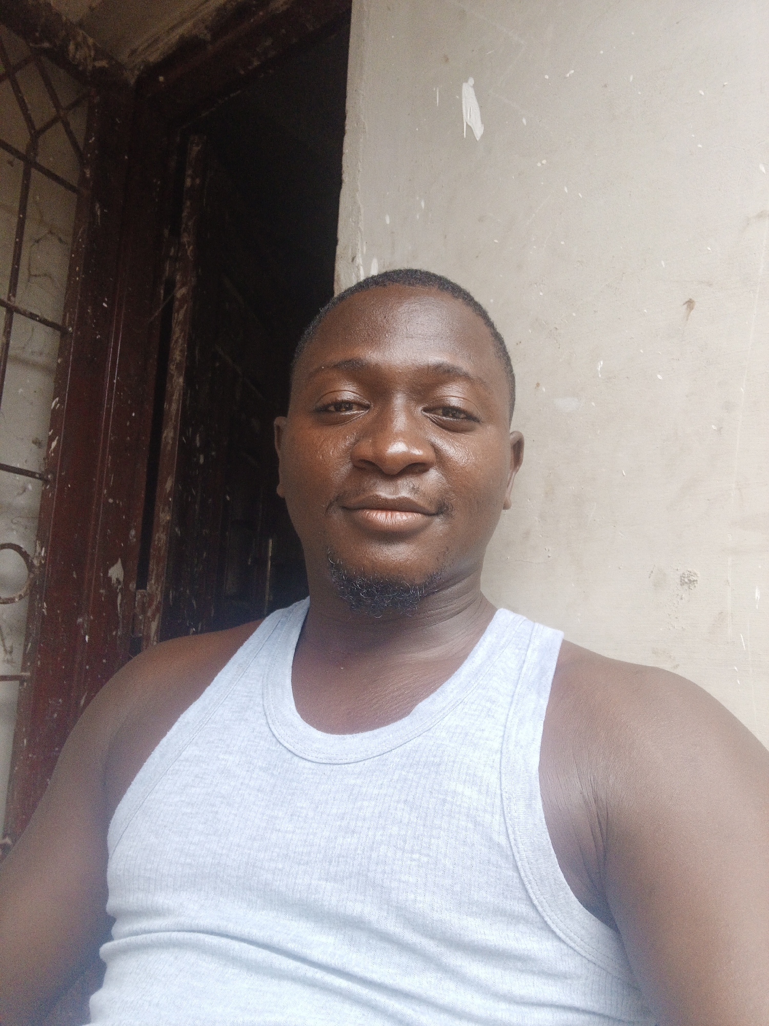 A man is captured in a close-up selfie, his face occupying the center of the frame. He has dark skin and short, dark hair, with a light stubble on his chin and jawline. He is wearing a light grey ribbed tank top. The man's expression is neutral, and he is looking directly at the camera. His eyes are dark, and his lips are slightly parted.
Behind him, on the left side of the image, is a dark, wooden structure that appears to be a door or window frame with metal bars. The wood is weathered and peeling, with visible signs of age and wear. To the right of the man, the background is a textured, light-colored wall, possibly concrete or stucco, with some splotches and marks on its surface. The lighting suggests it is daytime, with natural light illuminating the scene. The overall impression is a candid, informal snapshot taken outdoors or in an open doorway, possibly in a rustic or urban environment.