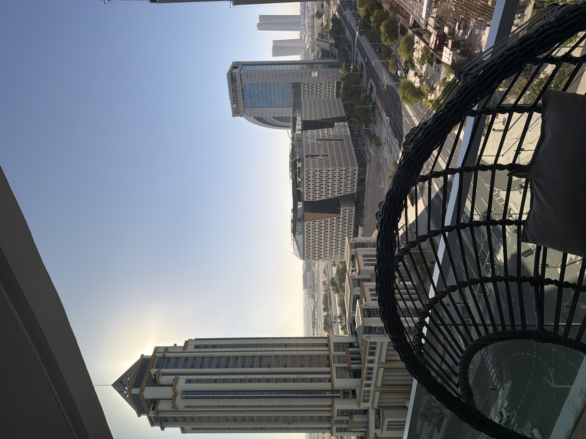 An outdoor, elevated, wide shot captures a city view under a clear, bright sky. The sun is partially obscured by a tall, ornate skyscraper on the left, casting a bright glare. In the middle ground, a modern, angular building with a patterned facade is visible, along with other contemporary high-rise structures. A road with a few cars is present in front of these buildings, bordered by trees and some construction activity. In the foreground, the top portion of a dark, woven wicker hanging chair with a pillow is in focus, obscuring the lower part of the scene. The overall impression is one of urban development and modern architecture.