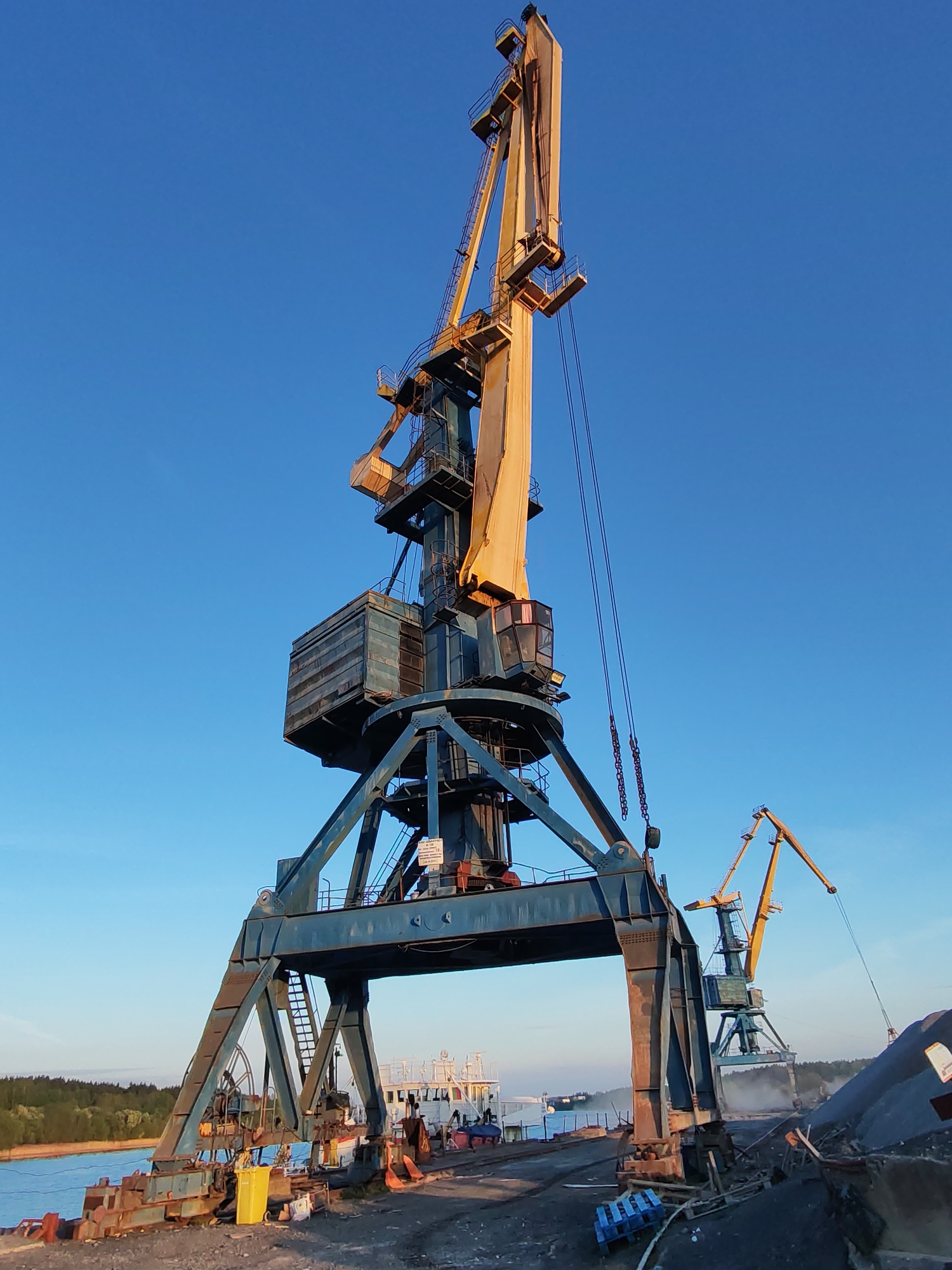 The image depicts two large harbor cranes in Tver, Russia. The primary crane is blue and yellow, towering over the scene. A second, similar crane is visible in the background to the right. The cranes are situated on a dock with evidence of industrial activity, including piles of materials and equipment. In the background between the legs of the crane is a small boat, likely a barge or tugboat. The sky is clear and blue, indicating daylight. The overall scene conveys an industrial setting at a river port.