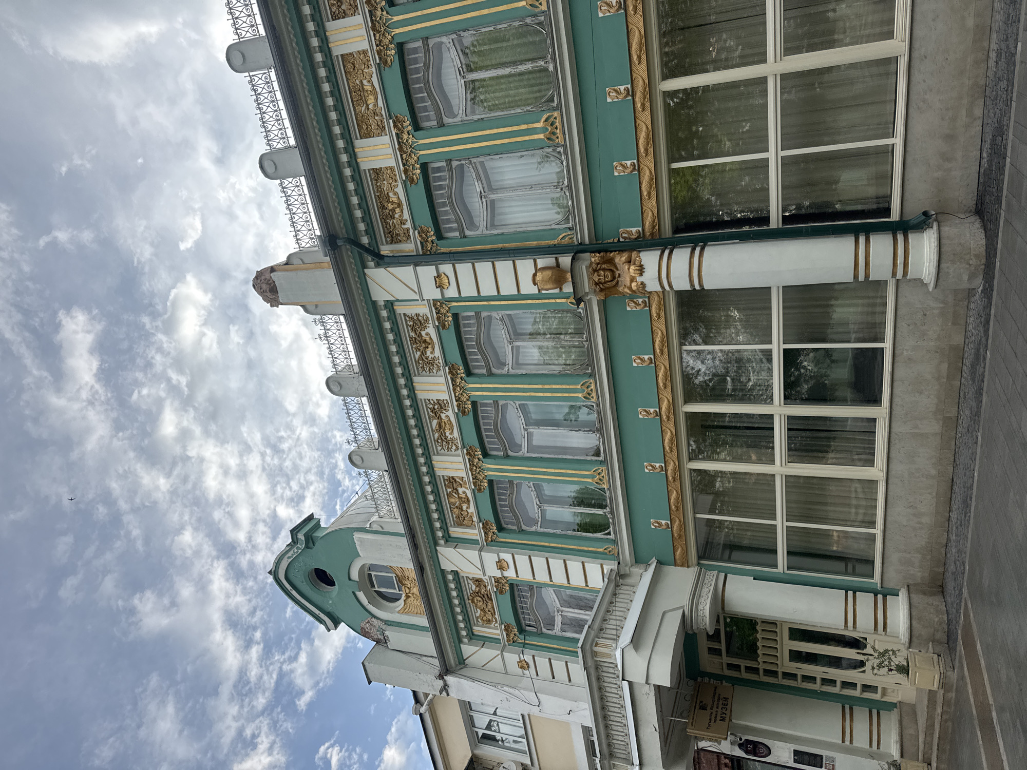 The image shows an ornate building in Vladikavkaz, Russia, with a green and white facade and gold detailing. The building is several stories tall with arched windows on the upper floors and rectangular windows on the lower levels. The facade is decorated with intricate gold patterns and sculptures. A wrought-iron railing is visible along the roofline. The sky is visible in the background, with scattered clouds.