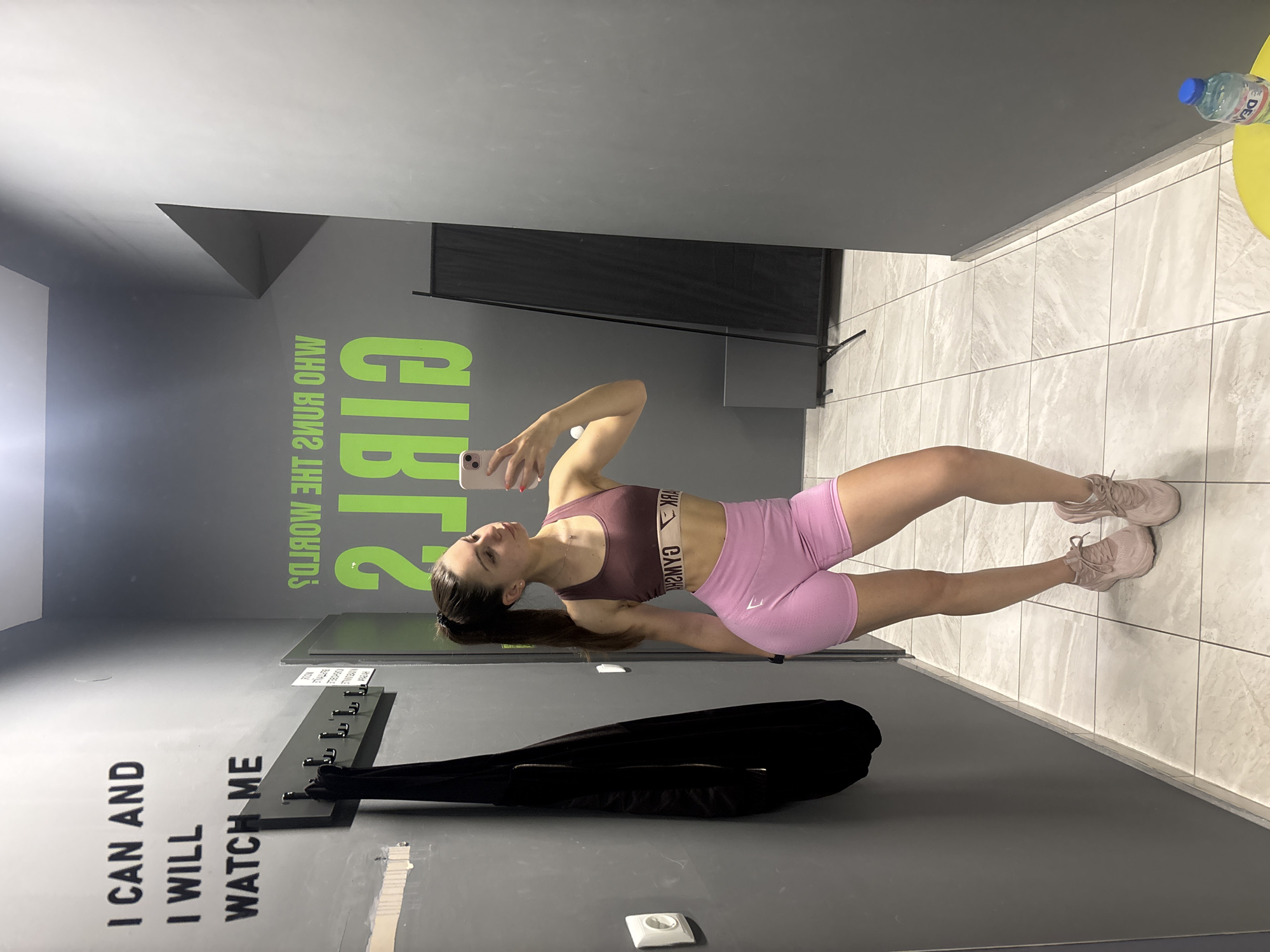 The image shows a young woman in a gym taking a selfie. She is wearing a burgundy sports bra and pink shorts and is holding a phone in her right hand. The gym has gray walls with motivational phrases like "I CAN AND I WILL WATCH ME" and "GIRLS WHO RUN THE WORLD." A black garment bag hangs on hooks on the wall. The floor is tiled in gray. A water bottle and yellow object rest in the background. The exact location within Varna, Bulgaria cannot be determined from the image.