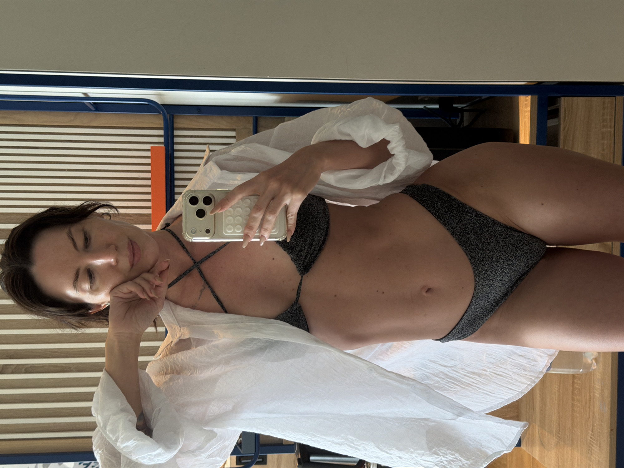 A woman with dark hair is taking a mirror selfie in what appears to be a hotel room. She is wearing a sparkly black bikini and an unbuttoned white sheer shirt. Her right arm is bent, with her hand resting on her cheek. She is holding a smartphone in her left hand to capture the photo. The background shows a wall with vertical wooden slats and a blue metal shelf. There is no indication of the city of Dong Ninh Hoa Ward, Vietnam.
