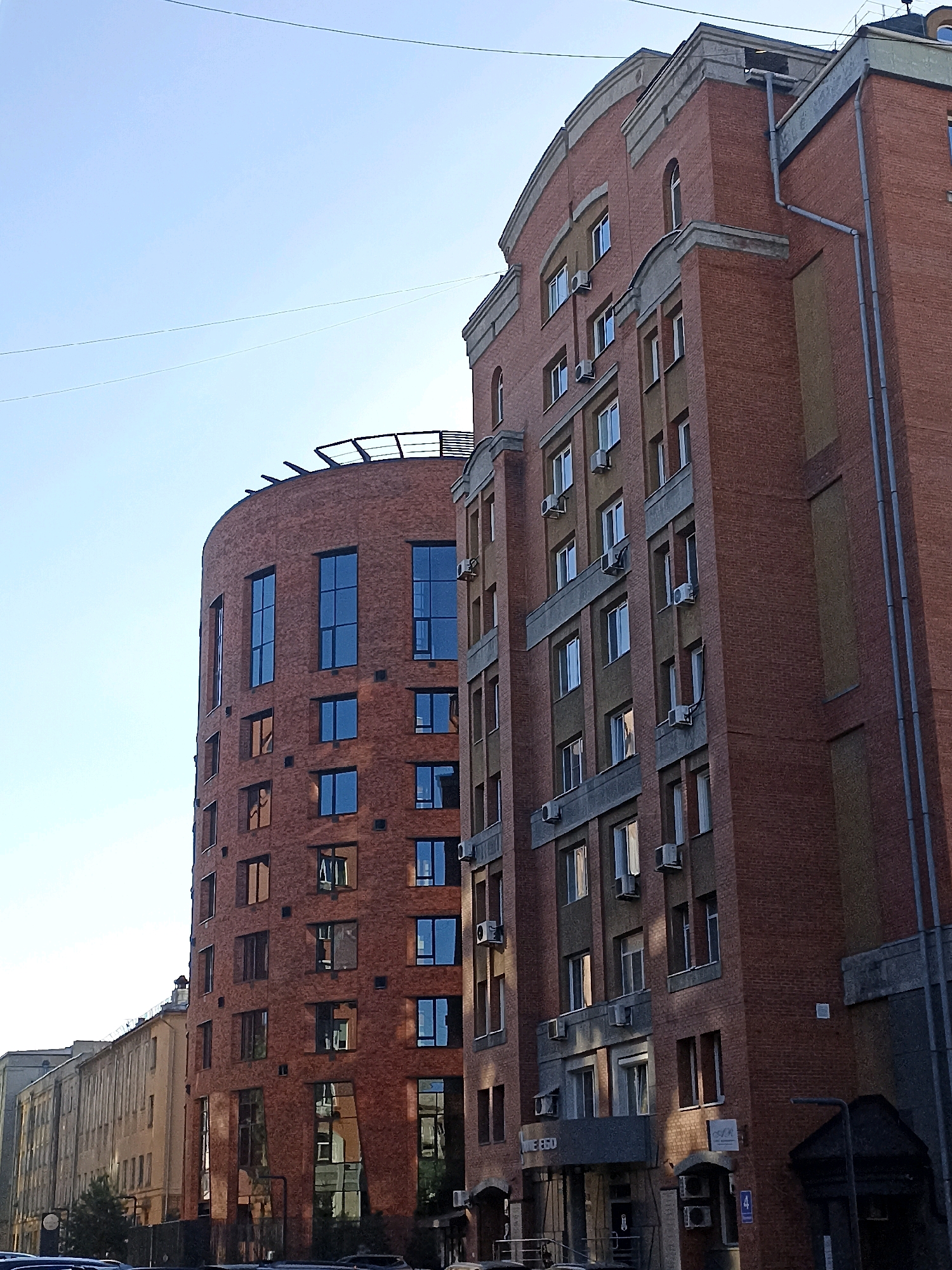 The image shows two modern brick buildings in Novosibirsk, Russia, on a clear day. The building on the right is taller and has a more traditional rectangular shape, with a red brick facade and multiple stories. It features numerous windows, some with visible air conditioning units installed outside. The building on the left is curved, with a distinct rounded facade also made of red brick. This building has larger, more stylized windows, some of which appear to be multi-pane. The sky is a clear, pale blue. In the foreground, a portion of a lower building and some trees are visible, suggesting an urban street setting. The overall impression is one of contemporary architecture in a city environment.