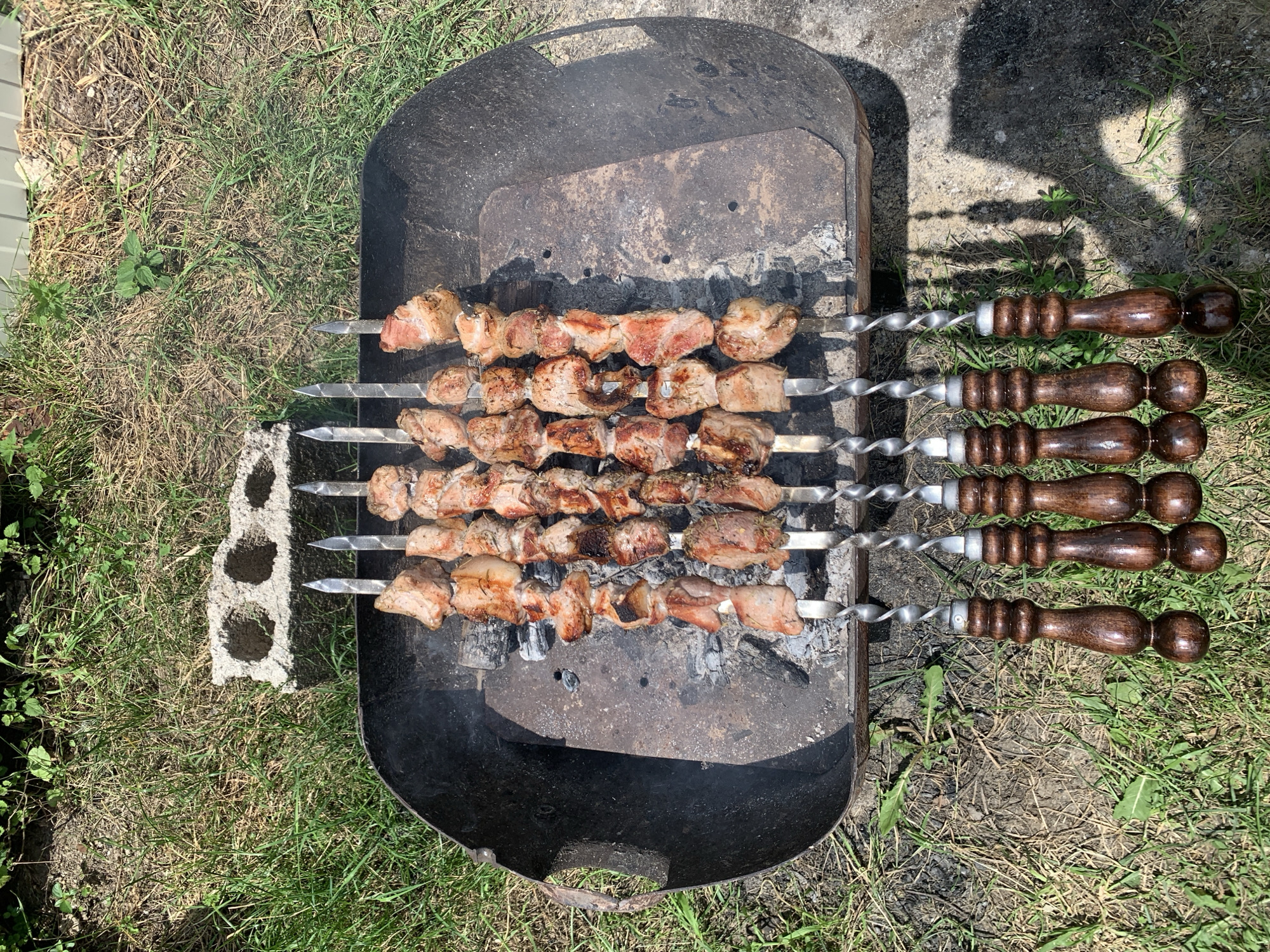 The image shows skewers of raw meat cooking over coals in a black metal barbecue grill. The grill is situated outdoors on a grassy area. There are several cinder blocks to the left of the grill and a concrete surface to the right. The skewers have decorative wooden handles. The meat is cut into uniform cubes and appears to be marinating. The sun is shining, casting shadows on the ground.