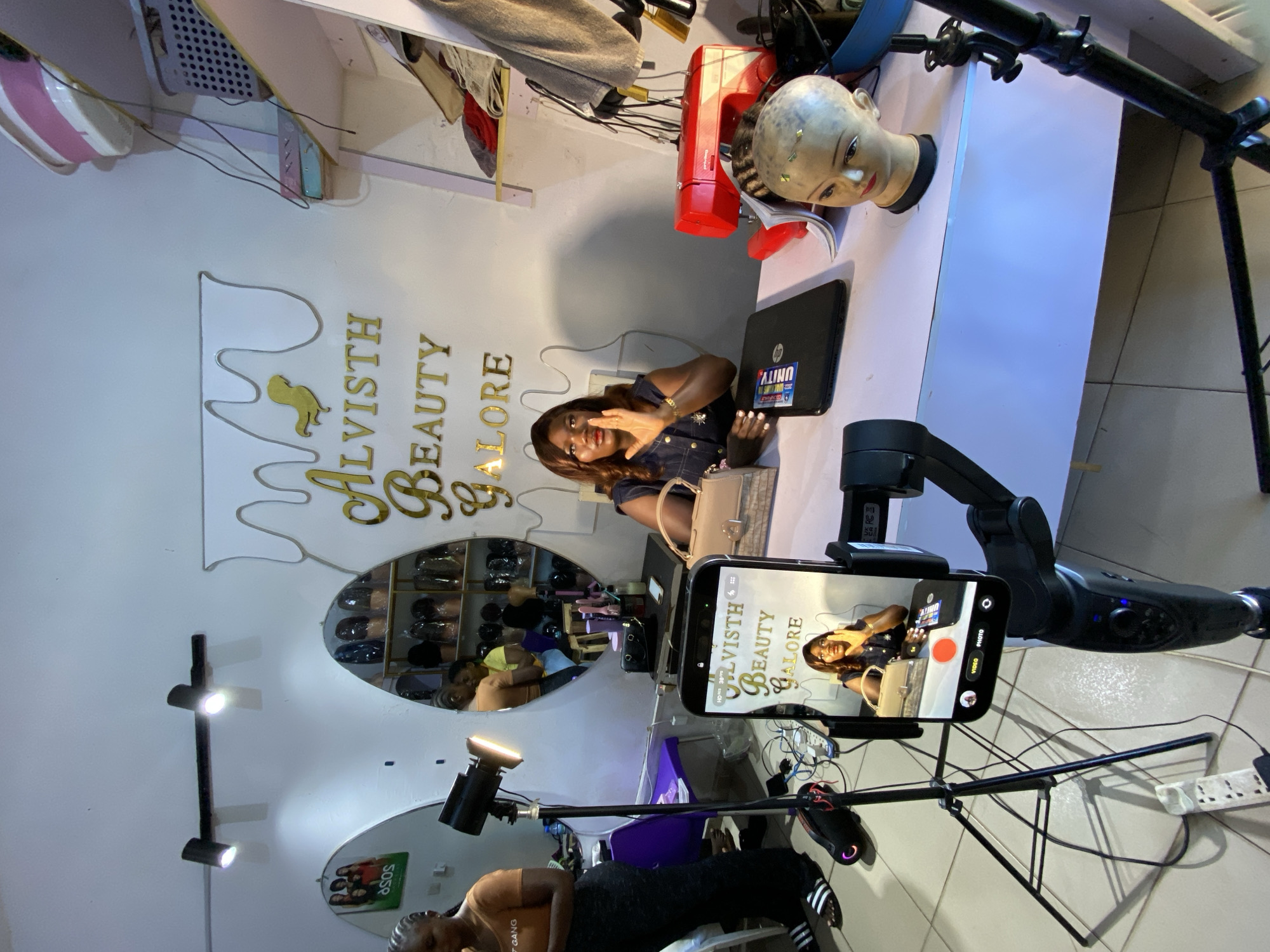 The image depicts the interior of a beauty salon, identified as "Alvisth Beauty Galore," located in Bukuru, Nigeria. The scene is set up for recording content, as indicated by a smartphone mounted on a gimbal, a ring light, and a professional tripod.
In the foreground, a woman is seated behind a white desk, facing the camera. She is wearing a dark denim dress and has her right hand raised, appearing to be speaking or gesturing. On the desk in front of her are a laptop with an "HP Unity" sticker, a red sewing machine, a mannequin head with hair, and a light brown handbag. The smartphone on the gimbal is capturing her image, displaying a live view of her and the salon's signage.
To the left of the frame, another woman, wearing an orange top and black pants, stands near a professional tripod with a ring light. She appears to be setting up or adjusting the equipment. In the background, a large mirror reflects shelves stocked with wigs and hair products, as well as another person partially visible. The walls are white, adorned with the gold lettering of the salon's name and a decorative dripping design. The overall environment suggests a functional and organized space dedicated to beauty services and content creation.