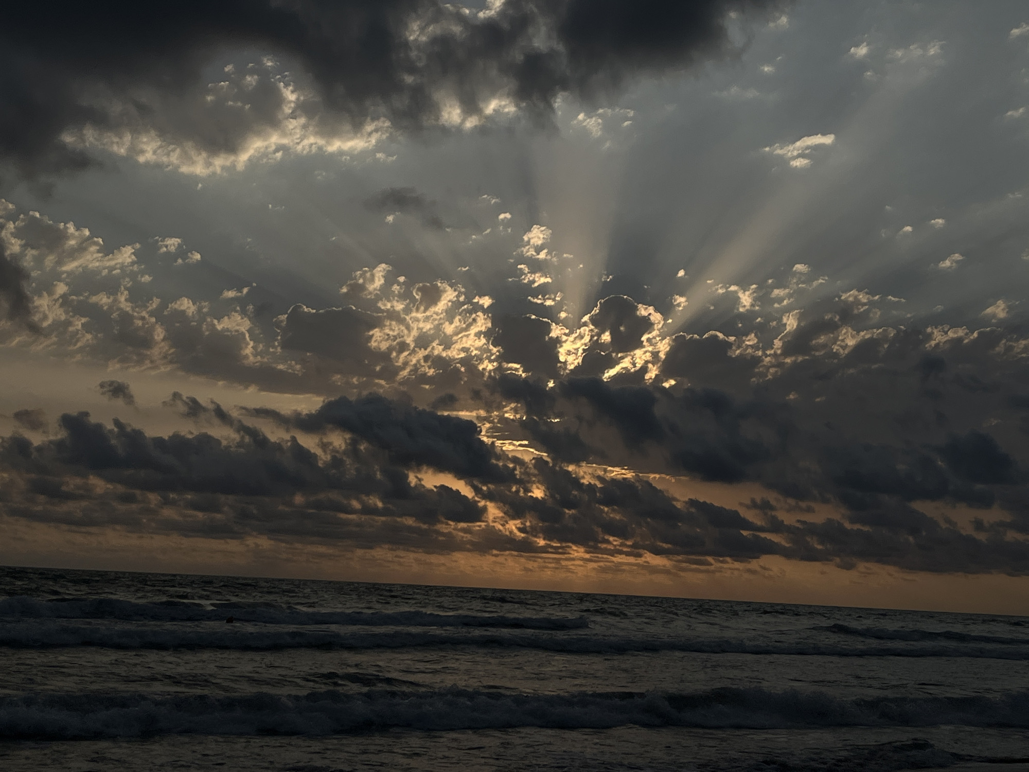 The image captures a dramatic sunset over the Black Sea, viewed from a beach in the Supasa community, Georgia. The sky is filled with a mix of dark, heavy clouds and lighter, illuminated clouds that are being pierced by rays of sunlight. The sun itself is not directly visible, but its light creates a radiant effect as it shines through the clouds, casting golden and orange hues across the sky. The sea is dark and choppy, with visible waves rolling towards the shore. The overall mood of the image is somber yet beautiful, with the powerful natural light contrasting with the dark storm clouds and turbulent sea. No people or man-made objects are prominently visible, emphasizing the raw power of nature.
