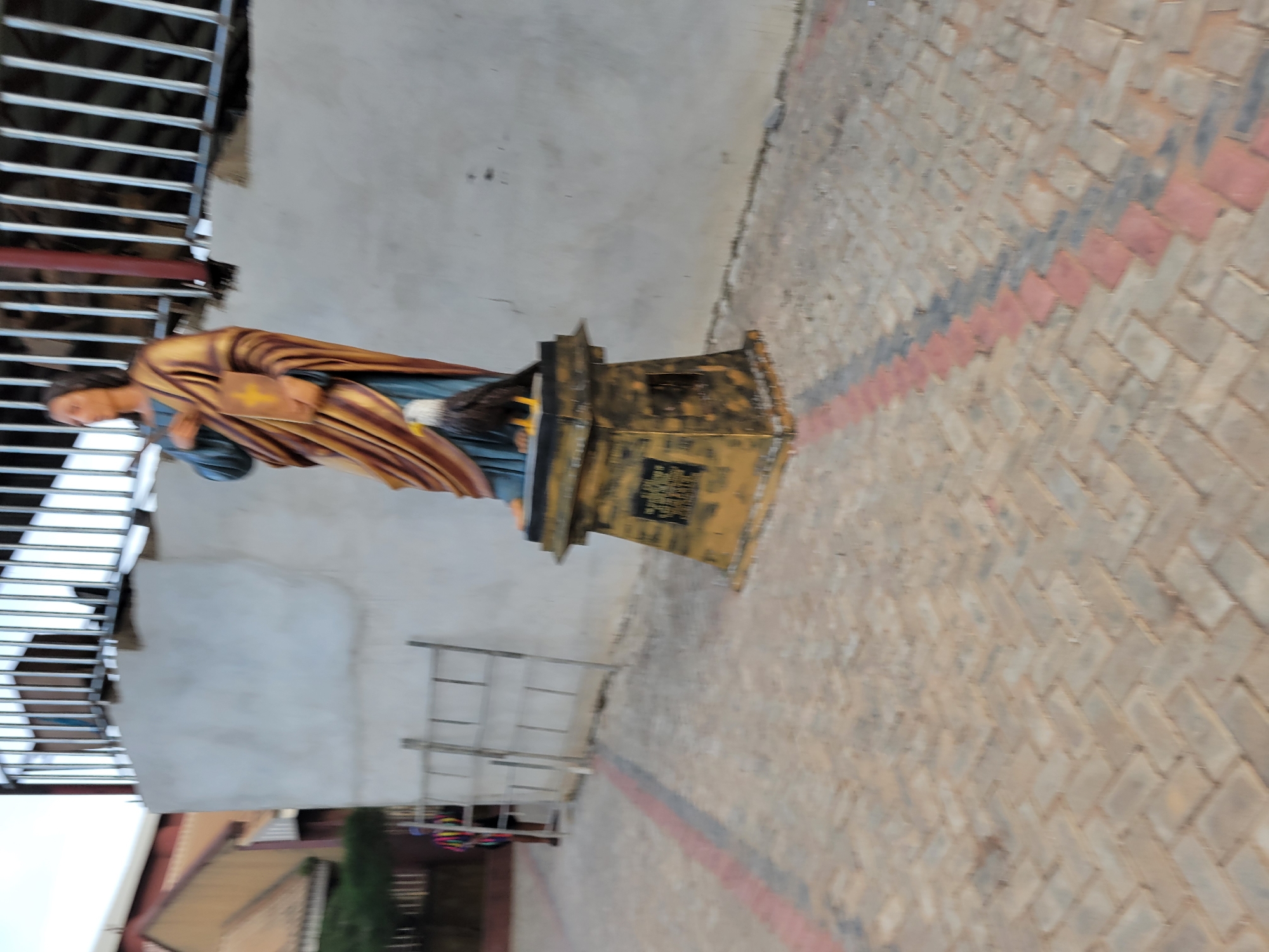The image shows a statue of a man holding a book, with an eagle perched on his arm. The statue is on a pedestal, which has some inscription on it. The background is a wall and a fence. To the left, there is a building with a red roof and a person in colorful clothing. The ground is paved with interlocking bricks in a herringbone pattern. The overall scene appears to be an outdoor religious or historical site. The location is identified as Onitsha, Nigeria.