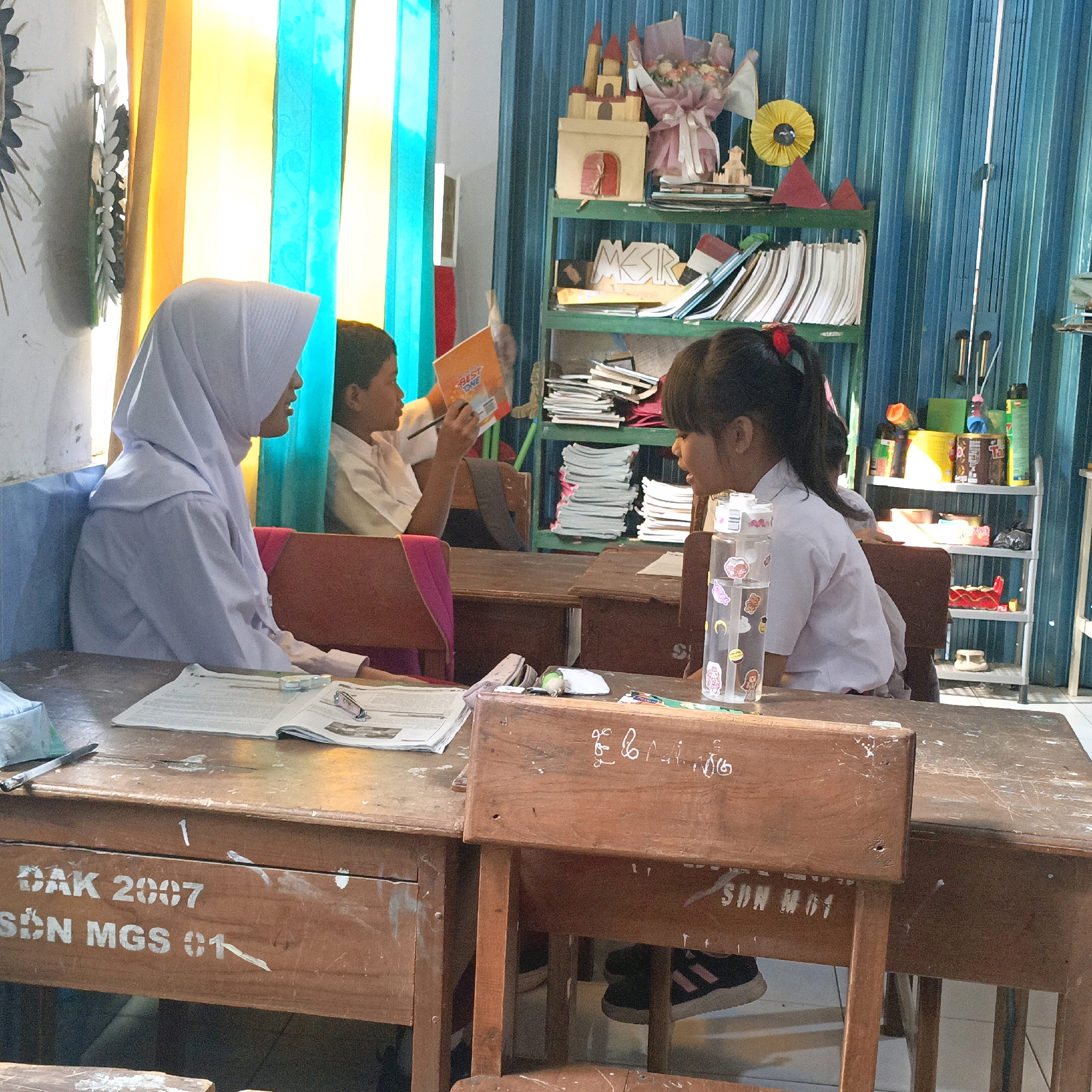 In a classroom in Salatiga, Indonesia, two young students are seated at desks. On the left, a girl wearing a light purple hijab and school uniform is looking down at an open book on her desk. To her right, a boy in a white school uniform is holding an orange book with the words "BEST ONE" visible and a pencil. He appears to be reading or studying. In the foreground, a wooden desk has an open book with a pair of glasses and a small stapler on it. On the side of this desk, painted text reads "DAK 2007 SDN MGS 01". On the right side of the image, another student, a girl with dark hair tied in a ponytail with a red ribbon, is wearing a white school uniform and looking to her right. A clear plastic water bottle with stickers adorns her desk. Behind the students, shelves are filled with books, notebooks, and decorative items, including a castle-like structure and a bouquet of flowers. The classroom is illuminated by natural light coming from behind bright yellow and turquoise curtains. The walls appear to be painted white and blue.