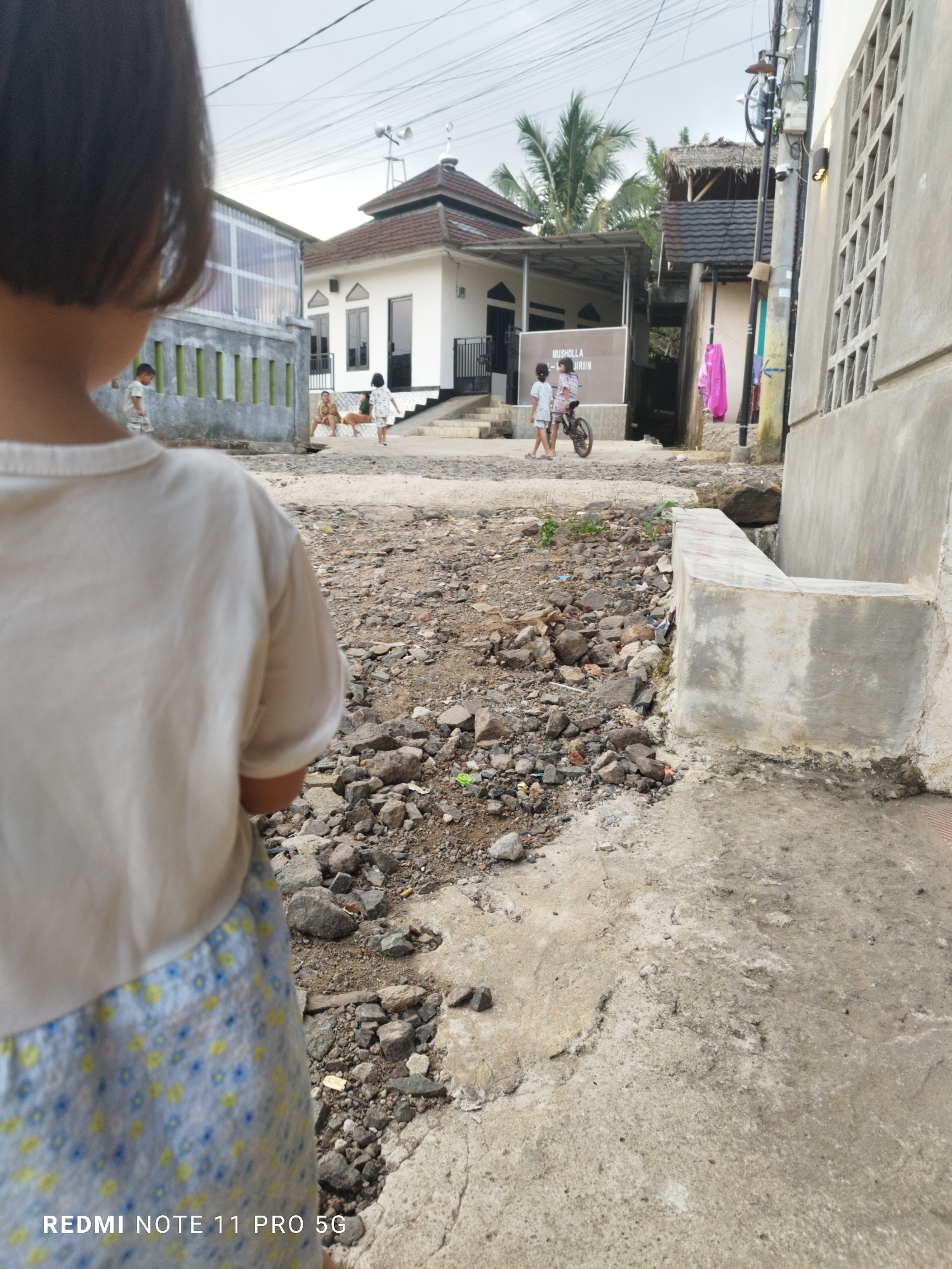 A child, seen from behind, stands in the foreground of a street scene in Kaduhejo, Indonesia. The child wears a light-colored t-shirt and a patterned skirt. In the background, several children are visible. Two children walk past a bicycle, and another two are seated on a raised concrete area. Behind them is a white building with a dark-tiled roof, resembling a mosque or musholla, with a sign that reads "MUSHOLLA AL-KHURIIN". A palm tree is visible behind a smaller building with a thatched roof. The ground is a mixture of dirt, gravel, and concrete. The sky is overcast.