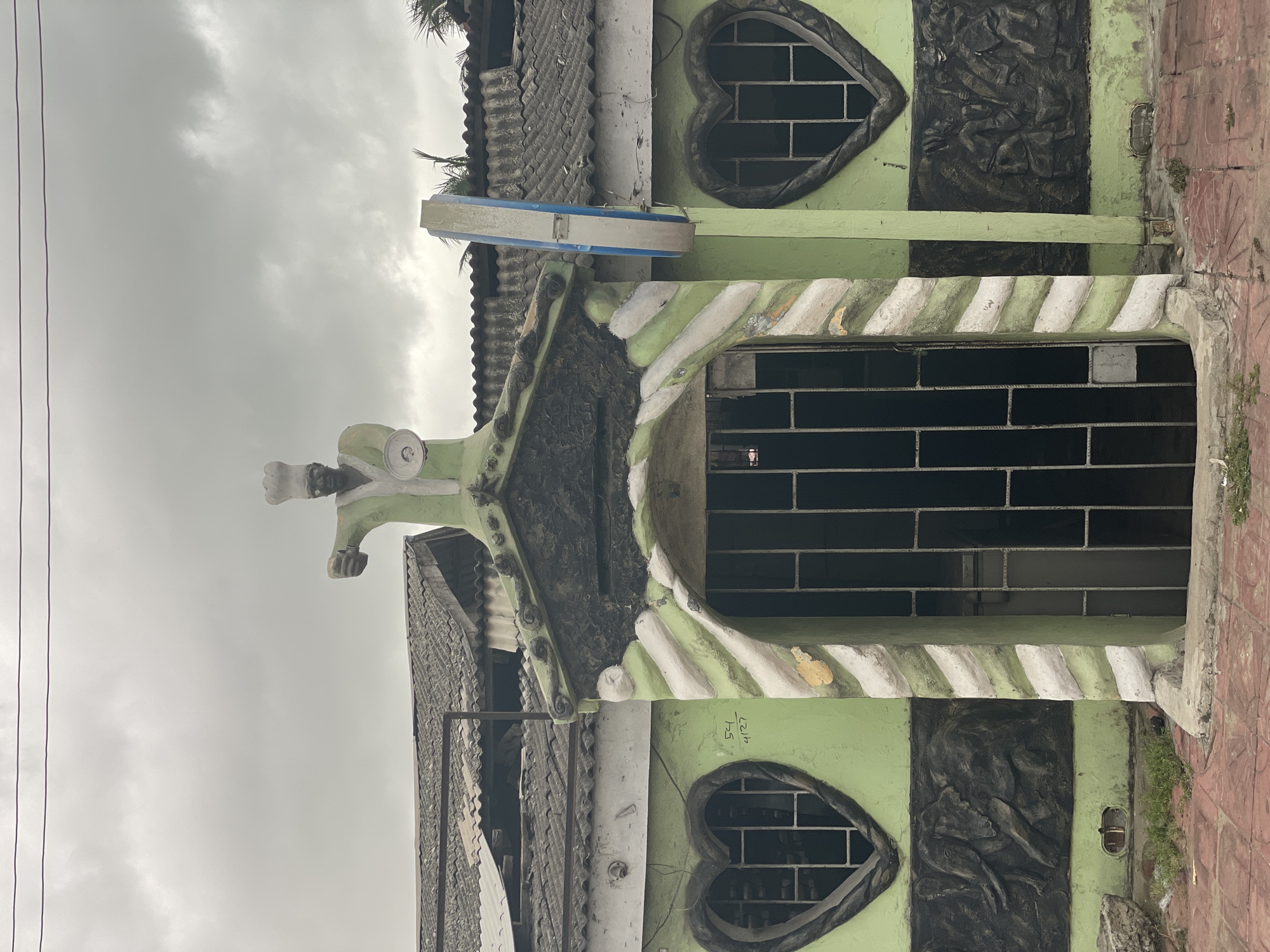 A building in Badagry, Nigeria, features a light green facade with two heart-shaped windows, both filled with a grid of black bars. Centered between the windows is a black, vertically-oriented relief sculpture. The entranceway, positioned between the heart-shaped windows, is similarly barred and framed with a pattern of alternating green and white diagonal stripes. The building is topped with a grey tiled roof, and additional sculptural details adorn the peak above the entrance. The sky is overcast.
