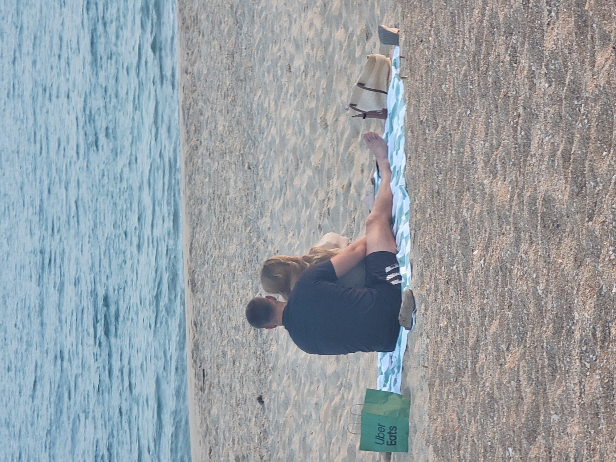 A couple is sitting on a blanket on the sandy beach in Quarteira, Portugal, facing the ocean. The man is wearing a dark blue t-shirt and black shorts, with his arm around the woman. The woman has blonde hair and is wearing a light-colored sweater. In front of them on the blanket is a green Uber Eats bag, a pair of sandals, and a straw tote bag. The ocean is calm and a light blue. The sand on the beach is light brown and textured.